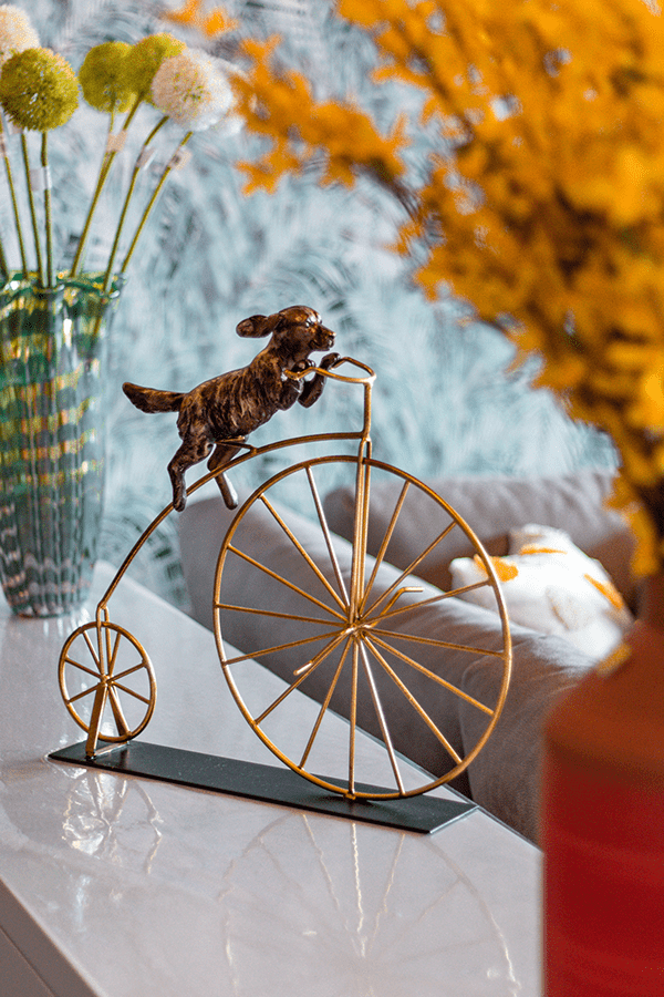 Decorative metal dog figurine riding vintage penny-farthing bicycle sculpture against autumnal background.