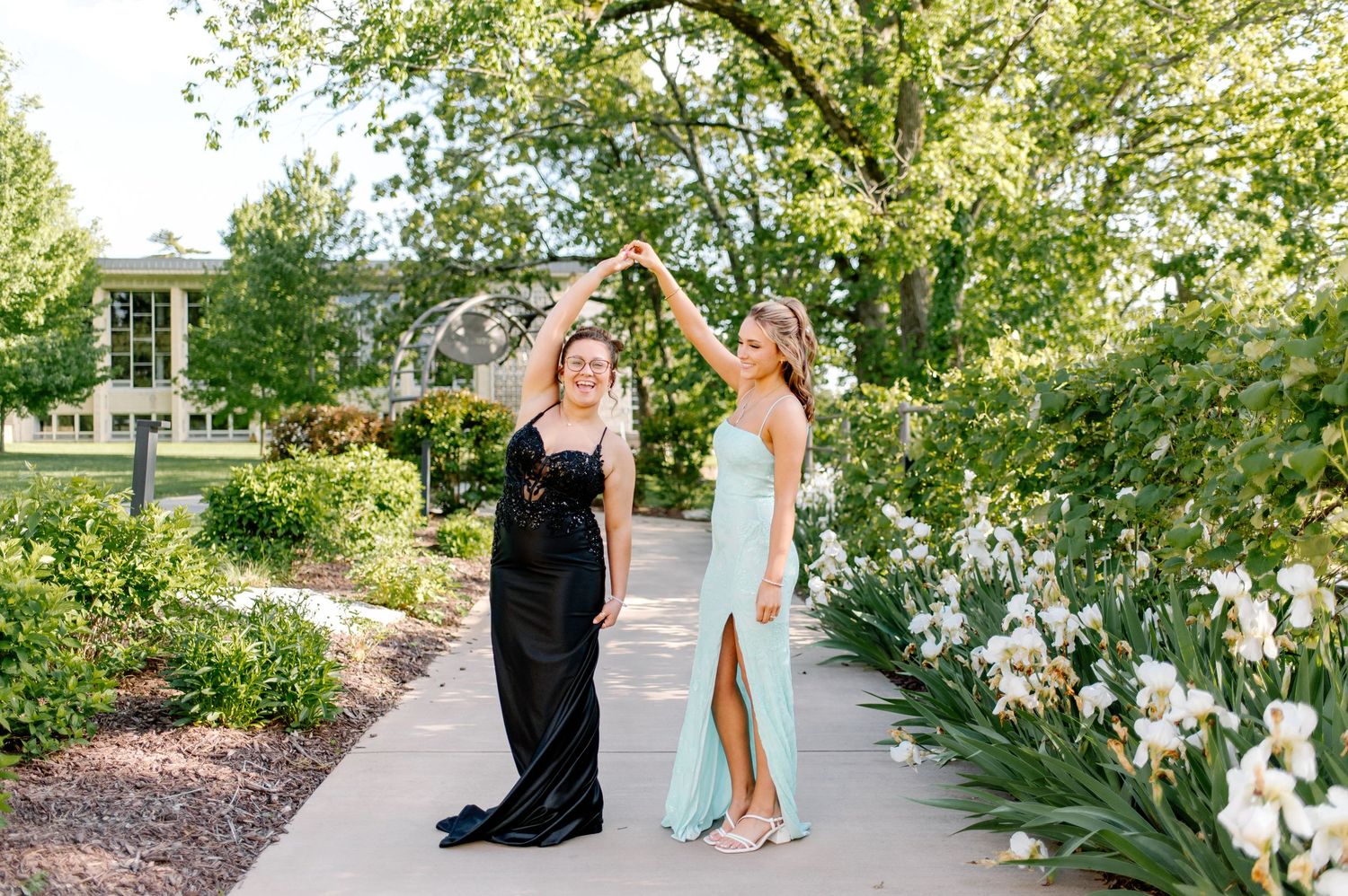 Wedding party walks along a garden path surrounded by lush greenery and white flowers on a sunny day.