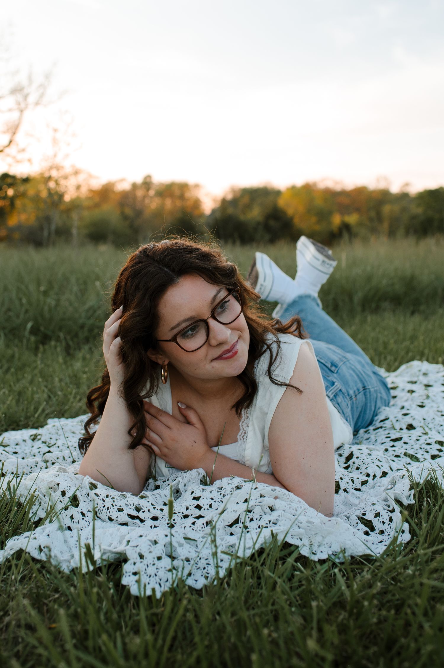 Person lying on white lace blanket in grassy field during golden hour sunset wearing glasses.