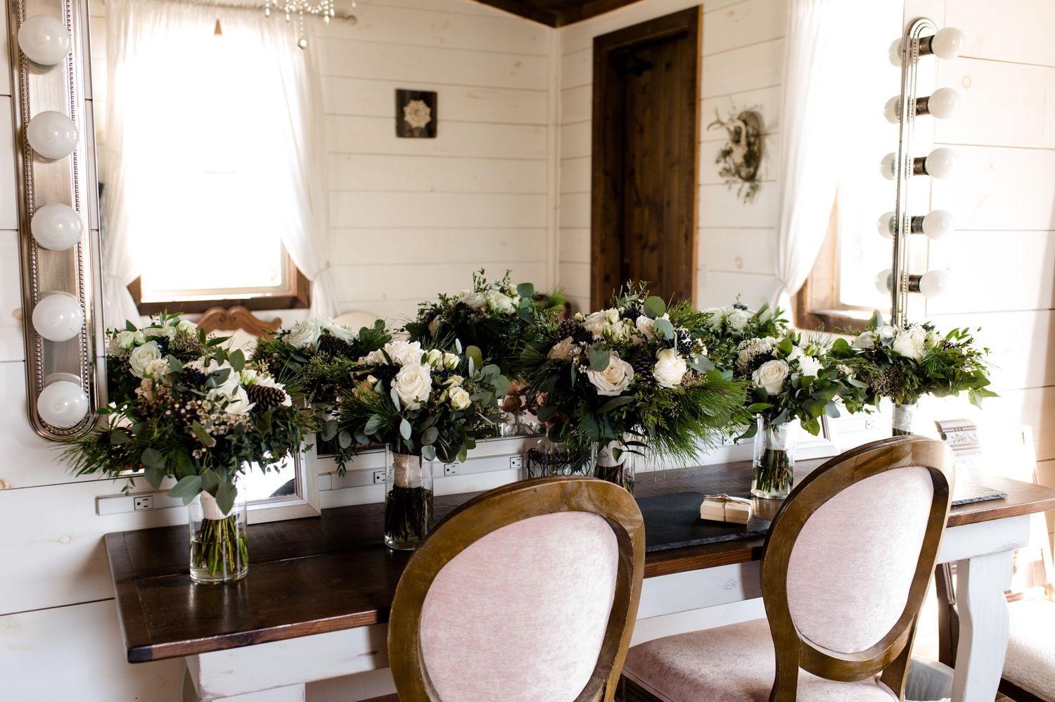 White floral arrangements with greenery decorate a wooden vanity table with vintage chairs.