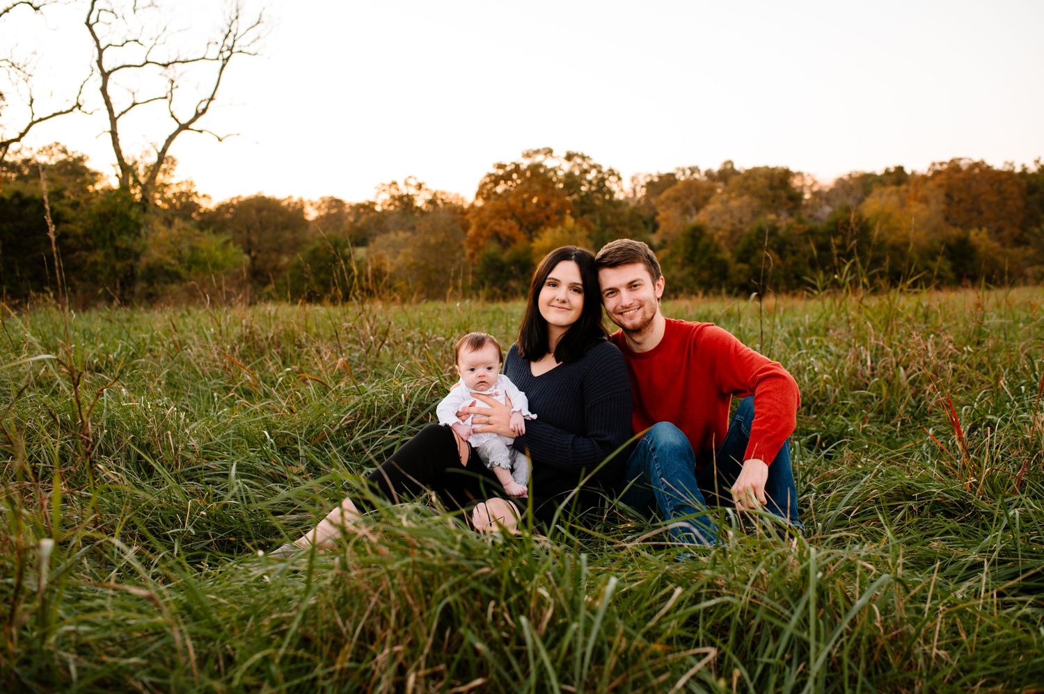 A family of three sits together in a tall grass field during sunset.