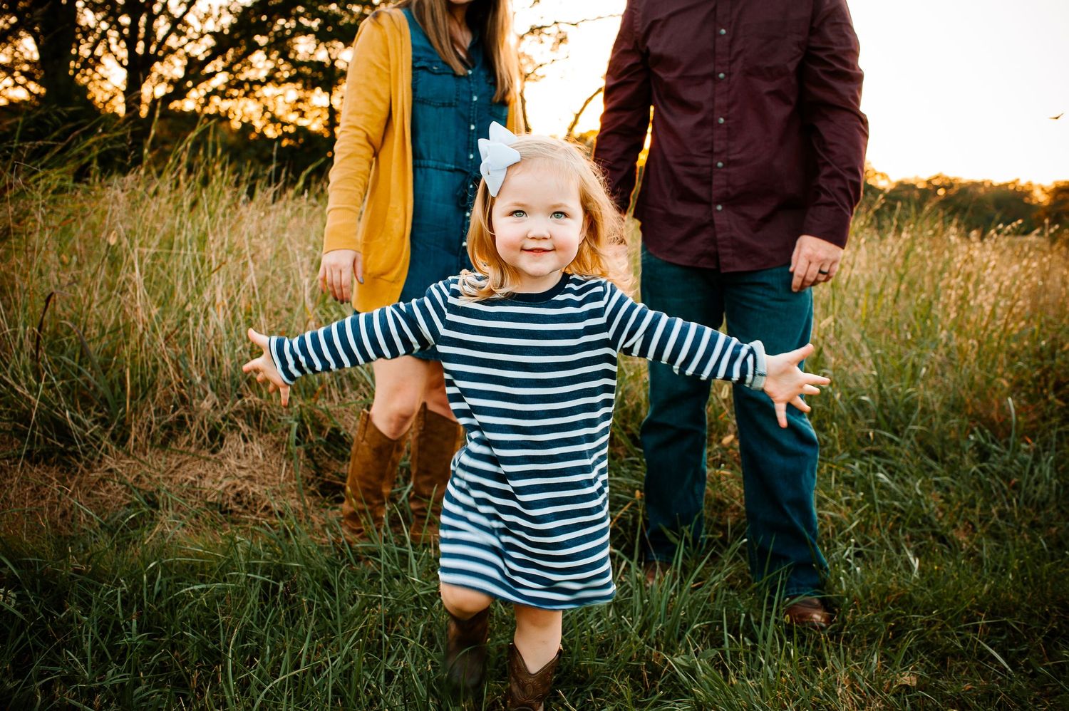 Child in striped dress running joyfully through tall grass during golden hour.
