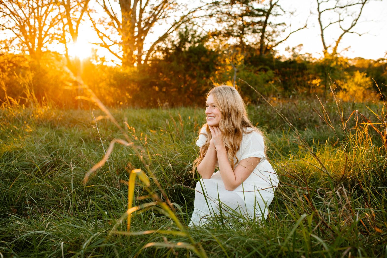 A person sits in tall grass during golden hour sunset with warm sunlight filtering through trees.