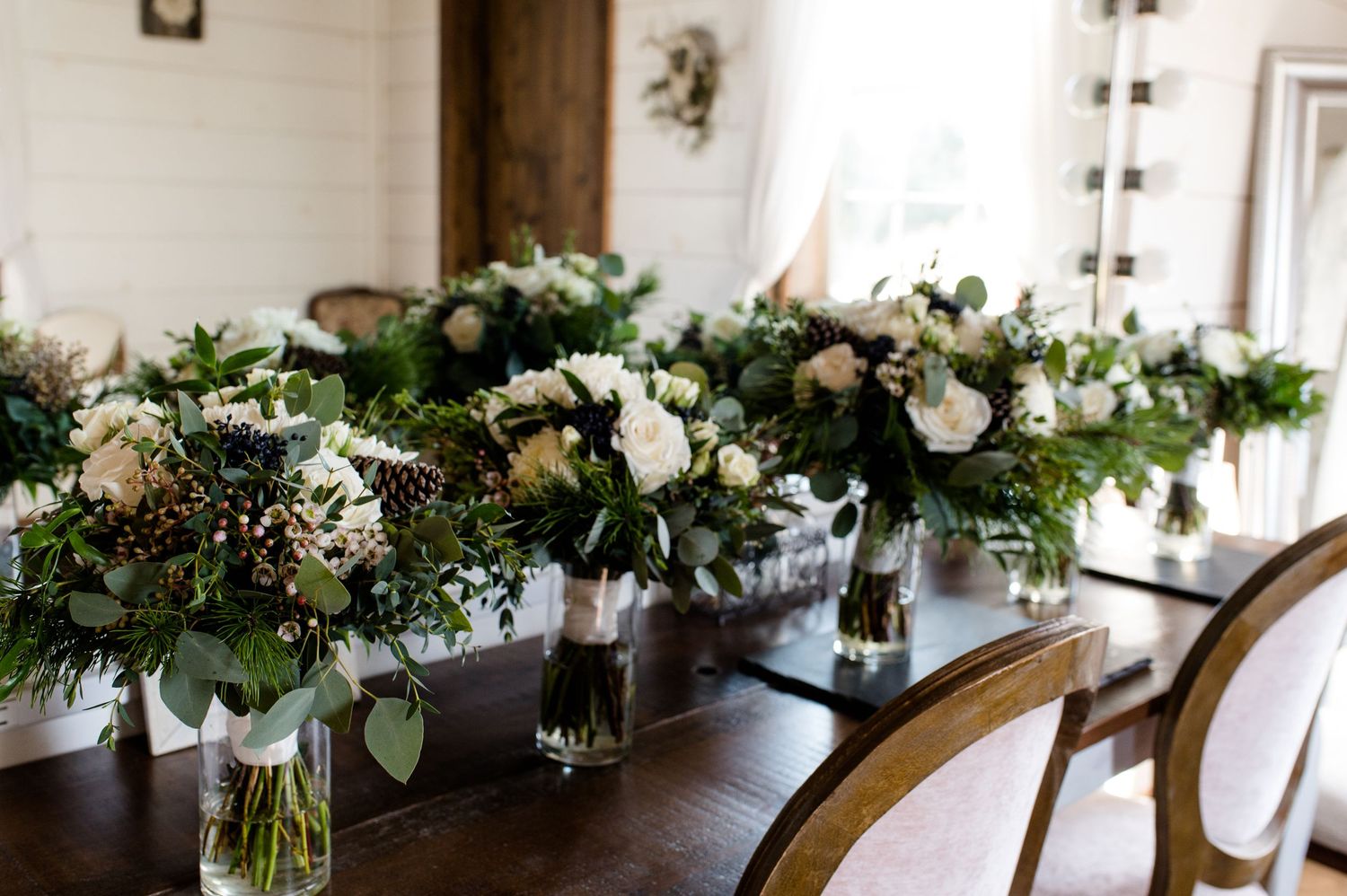 Elegant white floral centerpieces with greenery arranged on a wooden dining table for a wedding reception.