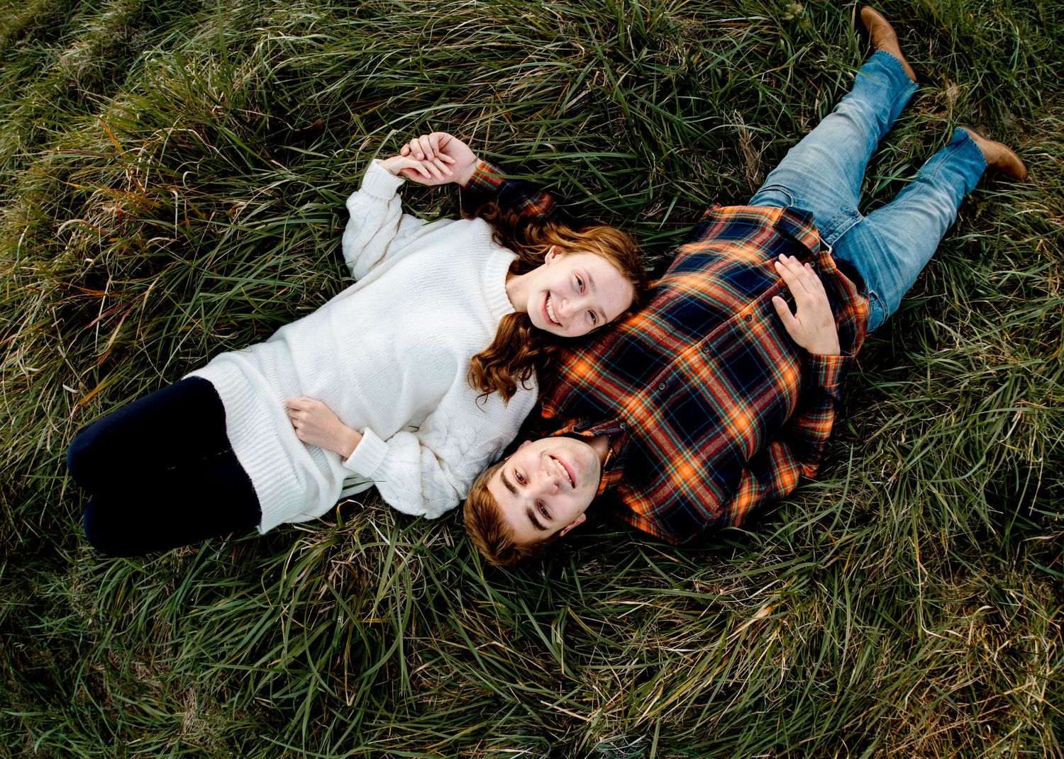 A couple lies in tall grass laughing together during an autumn photoshoot.