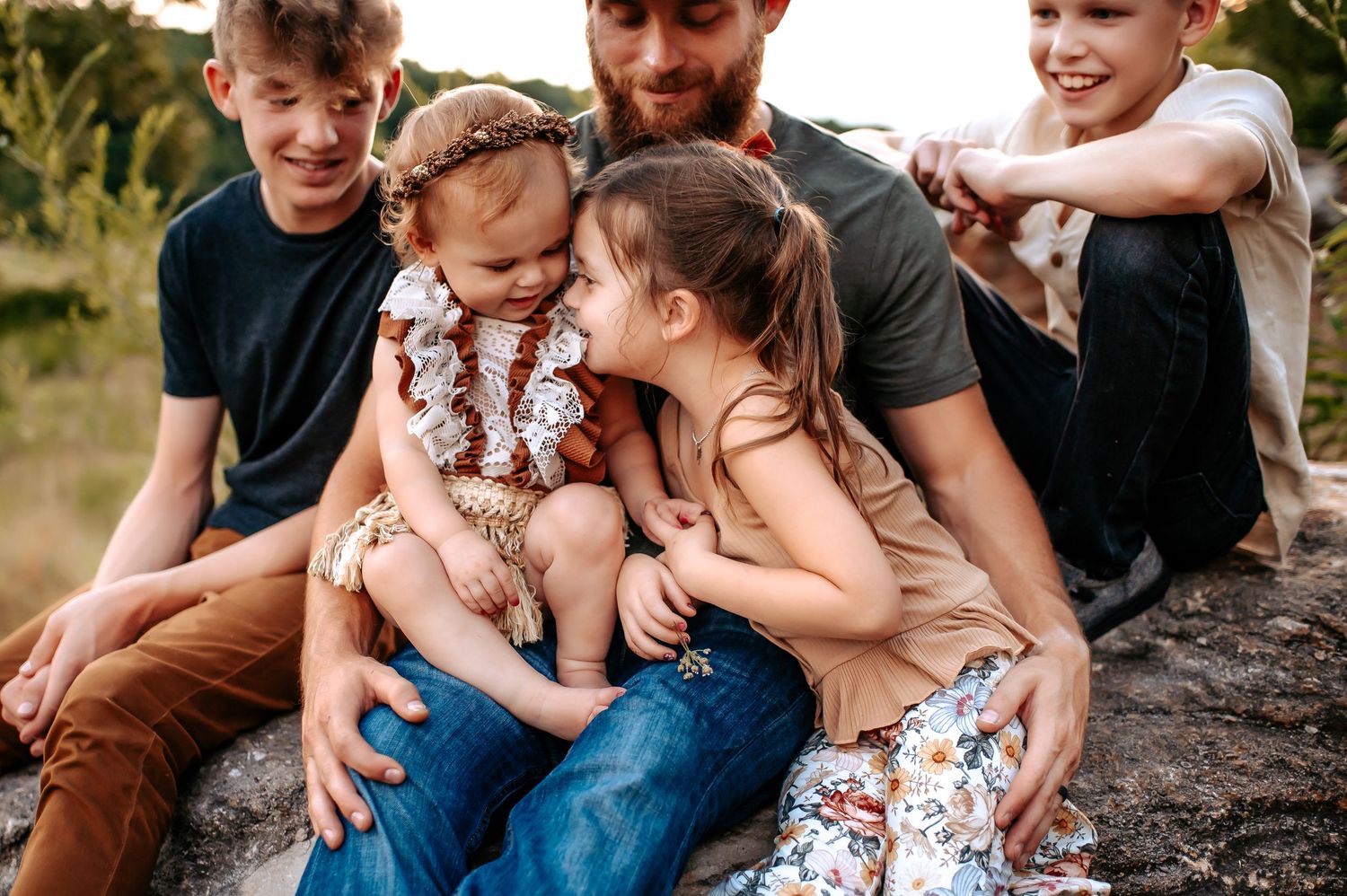 A candid family moment captured outdoors on a rock formation surrounded by natural foliage.