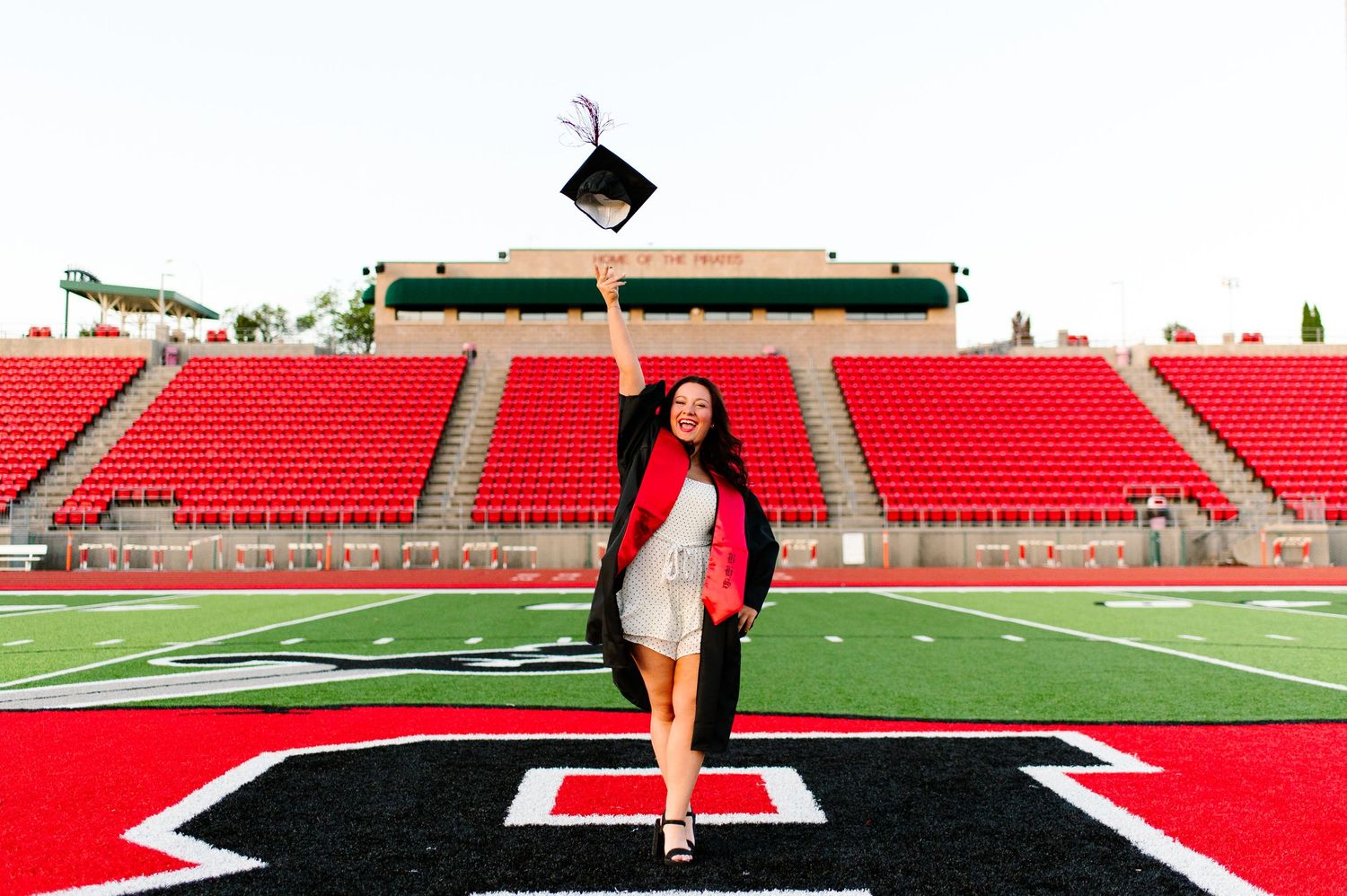 Graduate in cap and gown tosses mortarboard in stadium celebrating college graduation on football field.