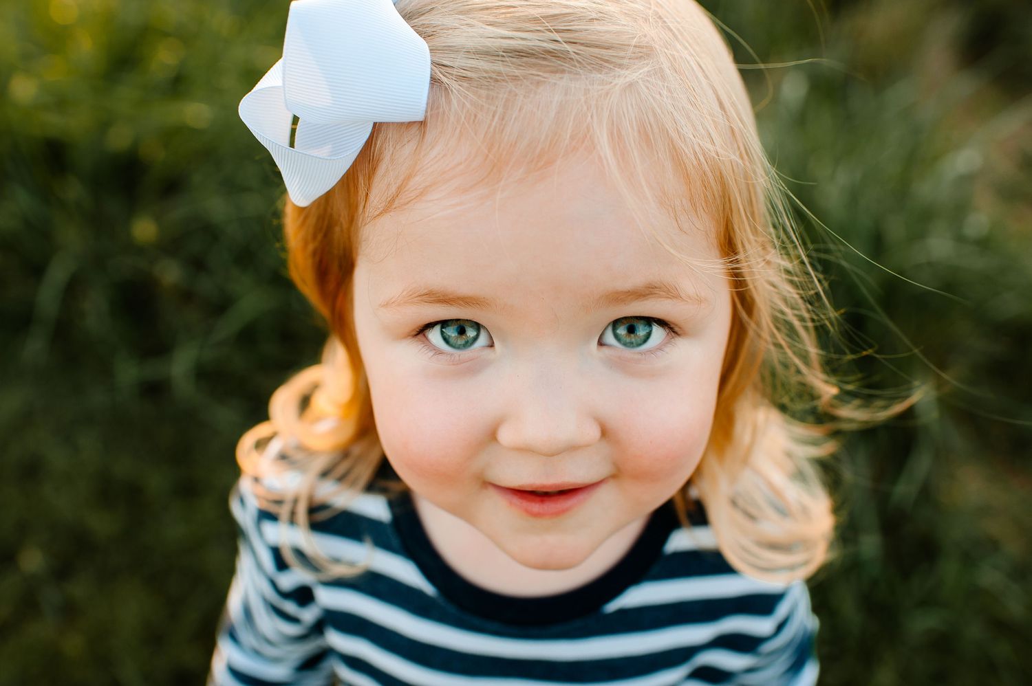 A close-up portrait of a young child wearing a striped shirt and white bow against a blurred green background.