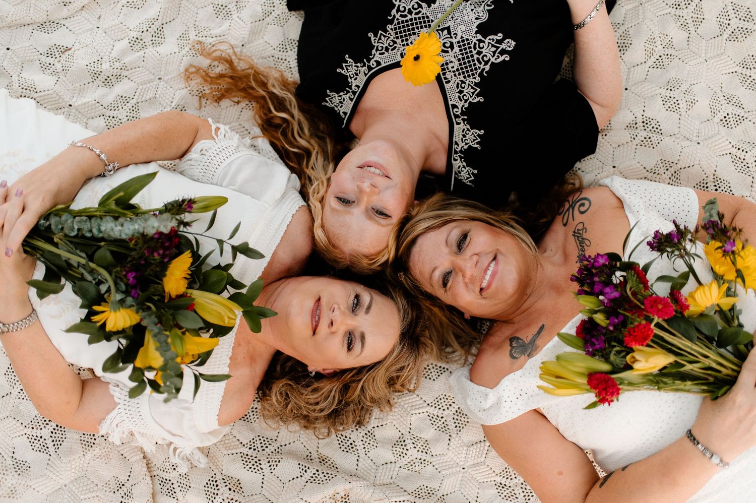 Three people lying on white fabric with colorful flower bouquets in an overhead shot.