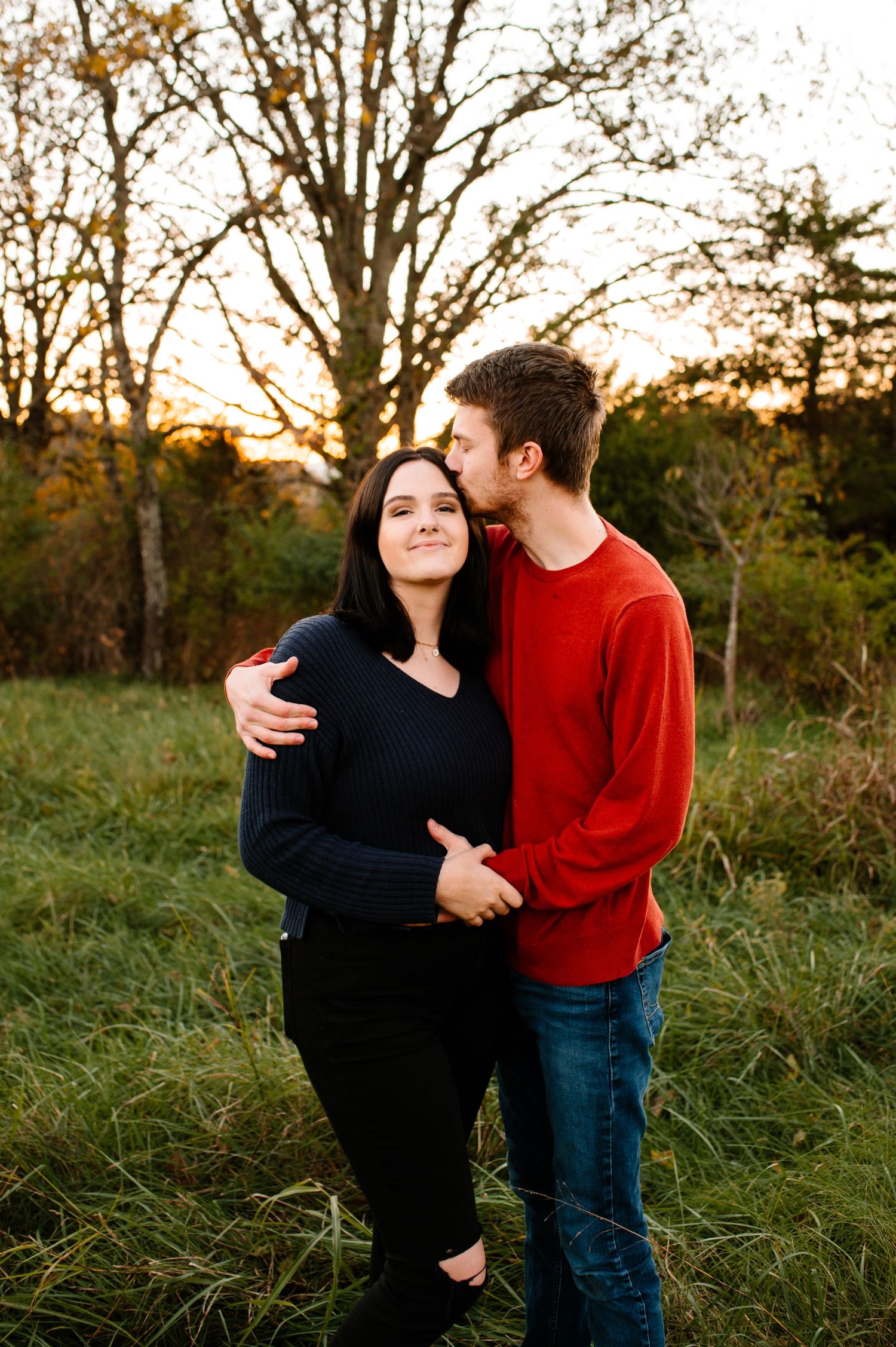 A romantic couple embraces in a field during golden hour with autumn trees in the background.