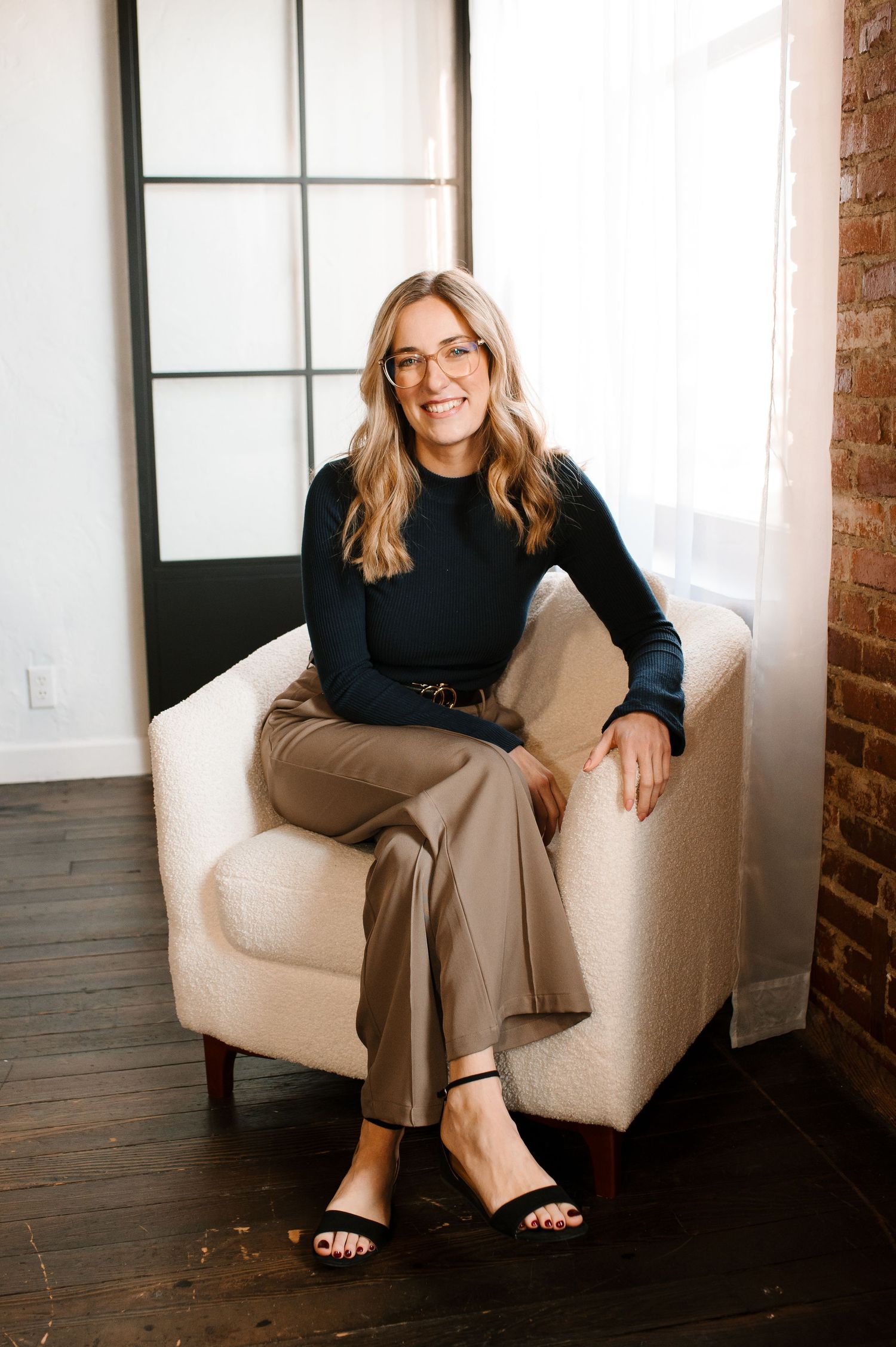 Smiling woman in black top and tan pants sitting in a white armchair in a modern studio space.