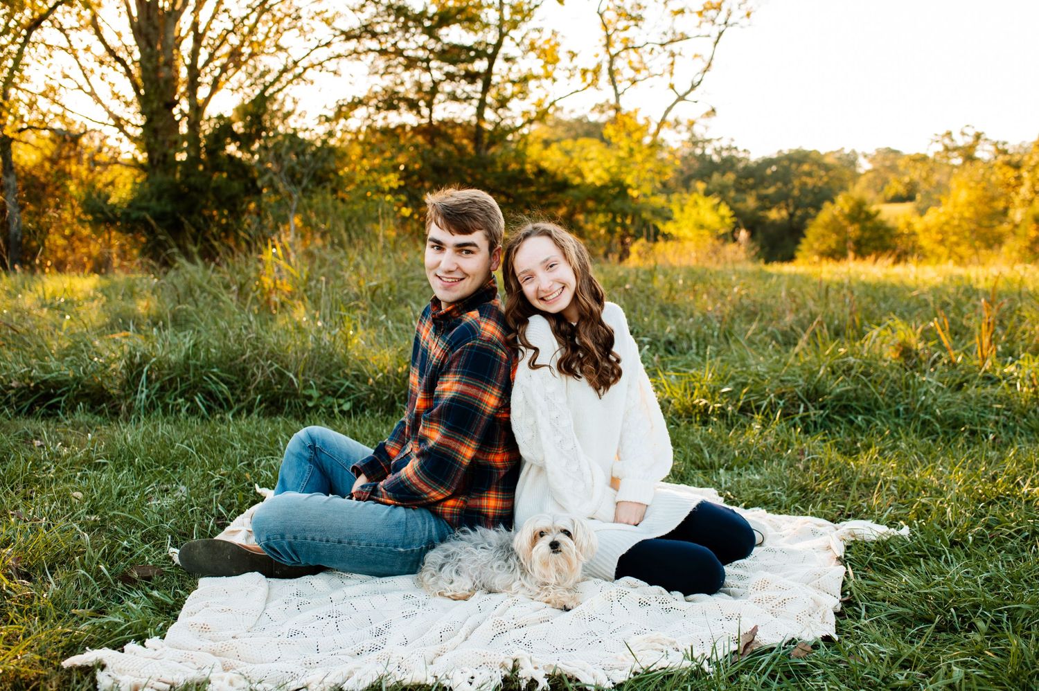 A couple sits with their small white dog on a blanket during a warm autumn evening photoshoot.
