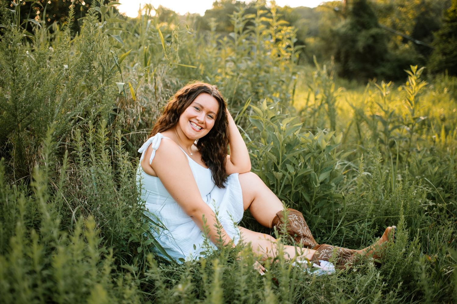 A person in a white dress poses gracefully among wildflowers and tall grass during golden hour sunset.