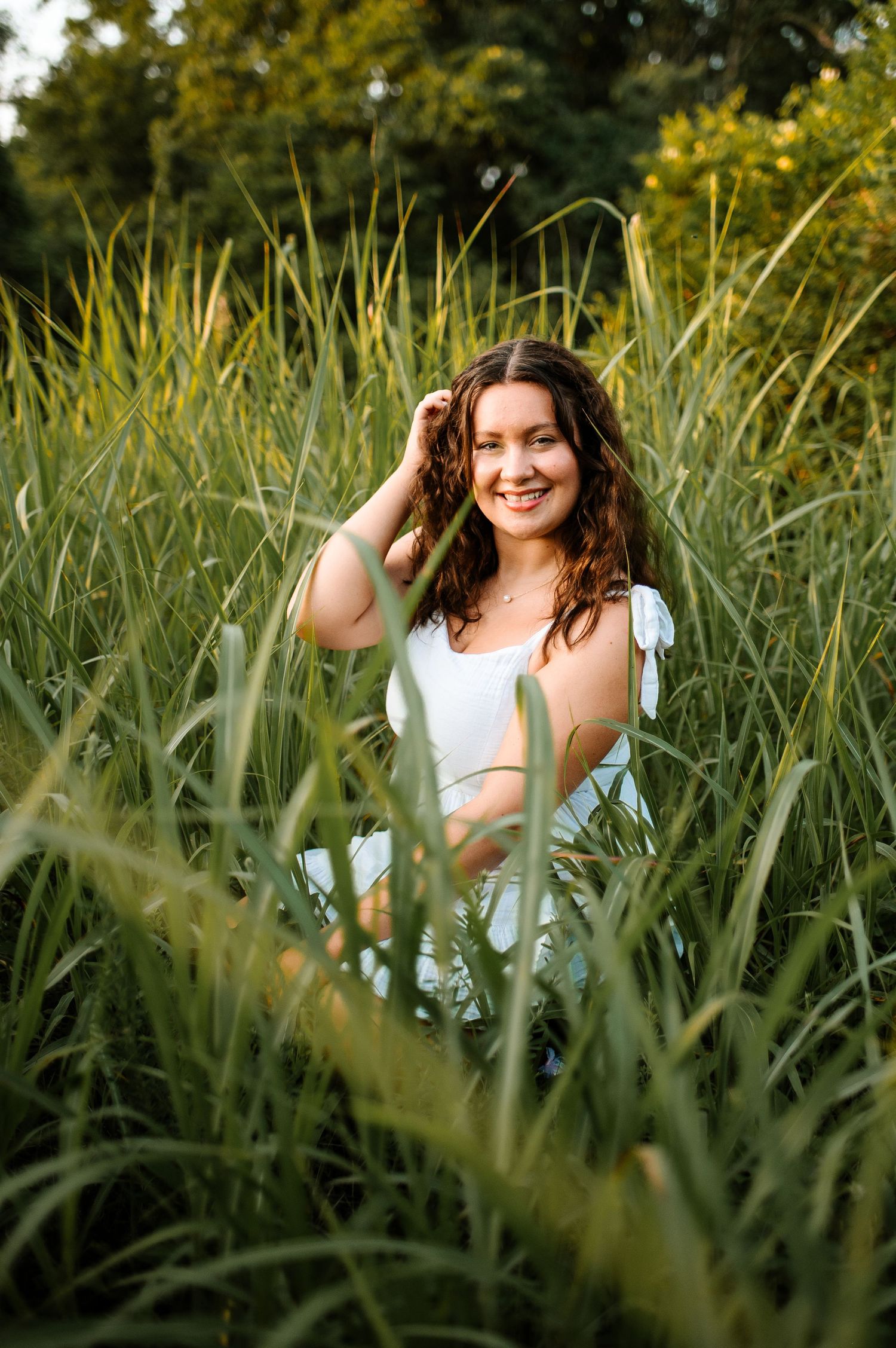 A woman in a white top poses among tall green grass and wildflowers on a sunny summer day.