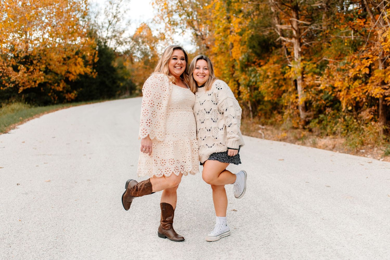 Two friends in white dresses playfully pose on a rural road during autumn.