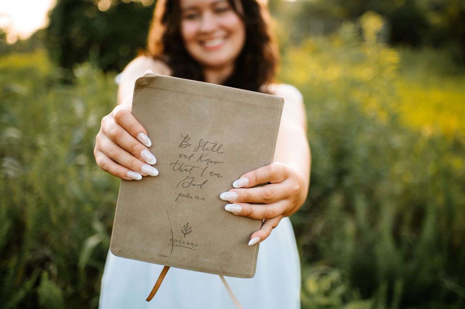 A person in a white dress holds a rustic brown envelope in a sunny meadow filled with yellow wildflowers.