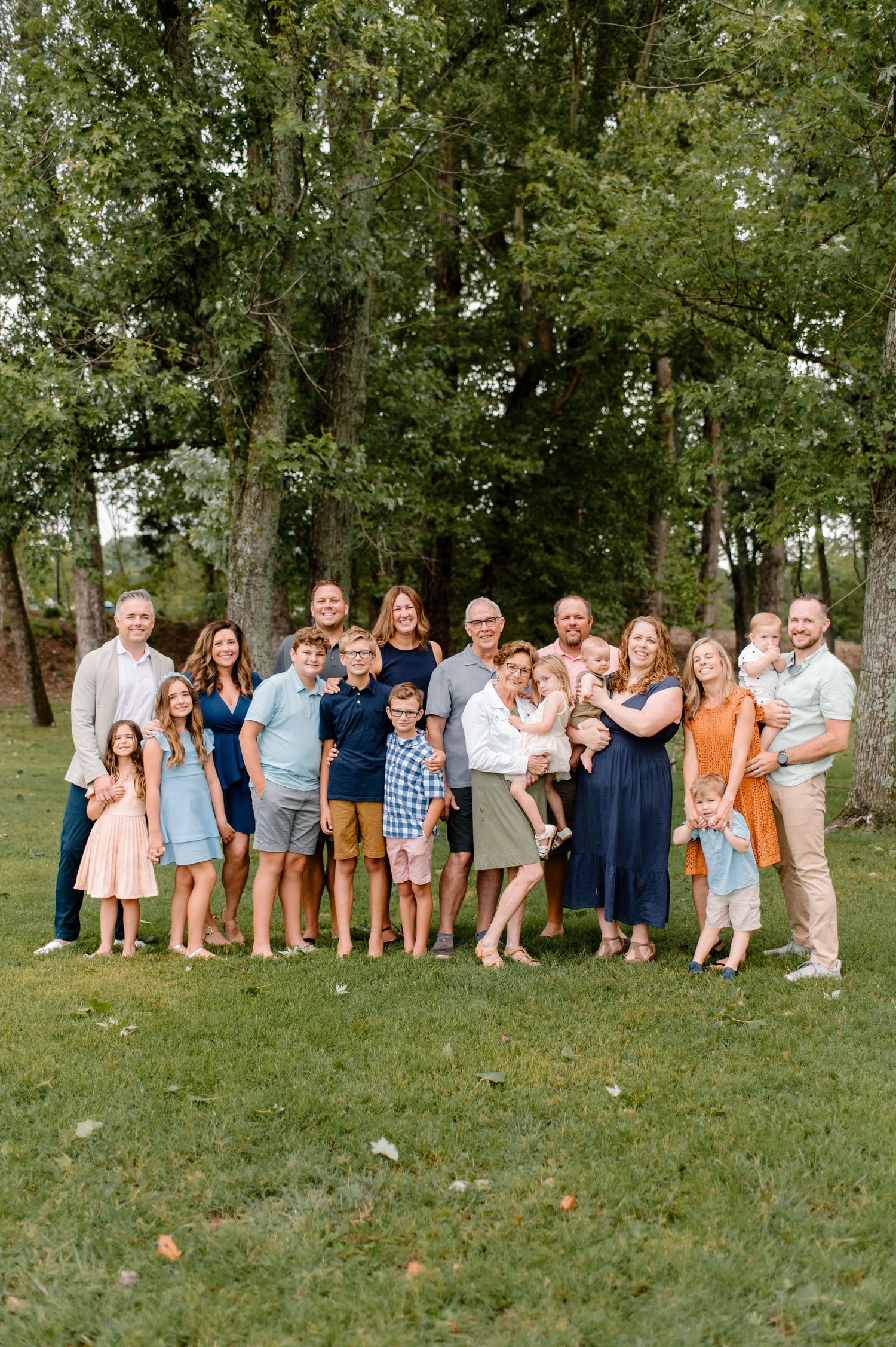 Large extended family group photo outdoors with coordinated casual outfits against green trees.