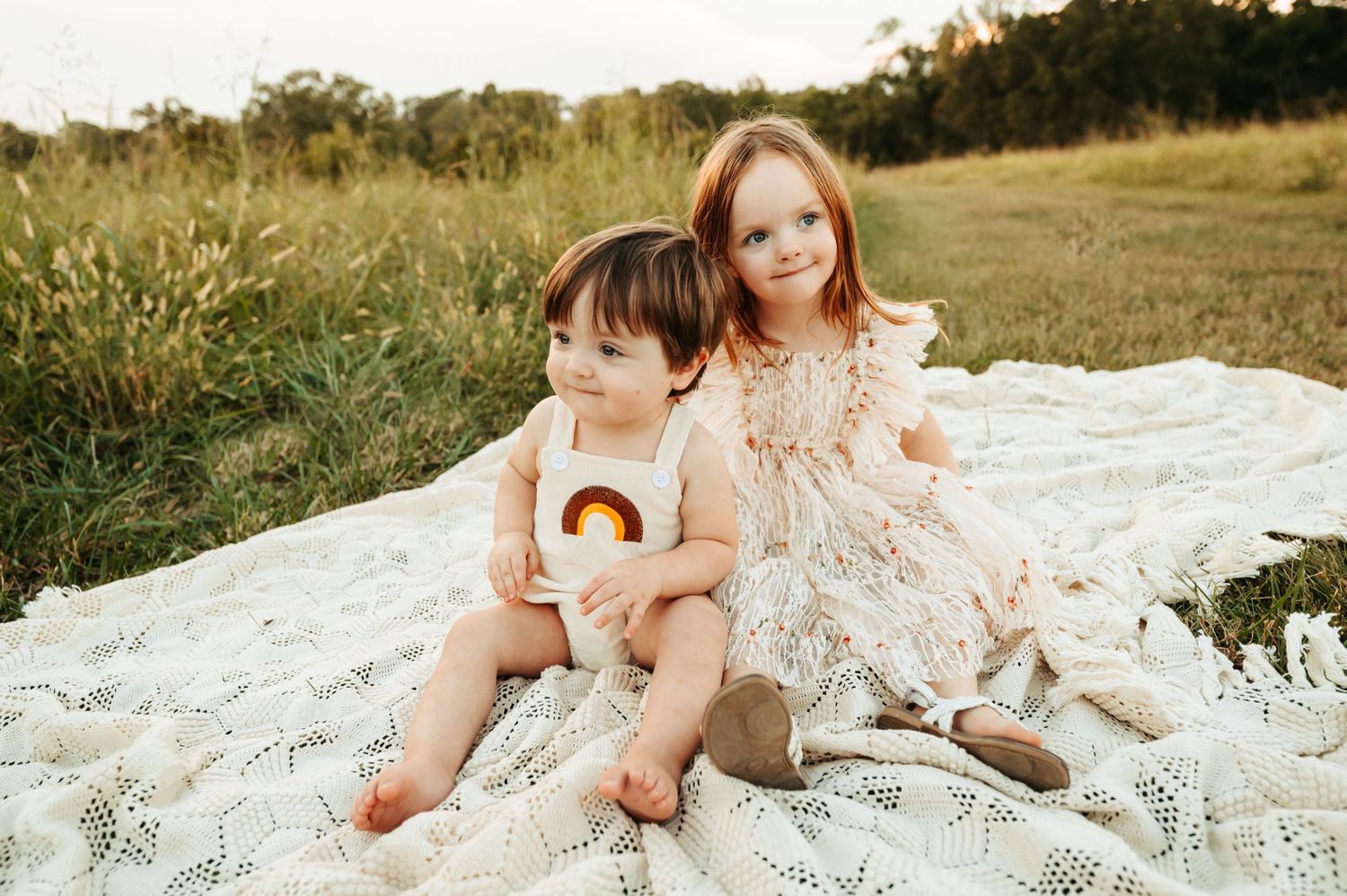 Two toddlers sit together on a white blanket in a grassy field at sunset wearing neutral colored outfits.