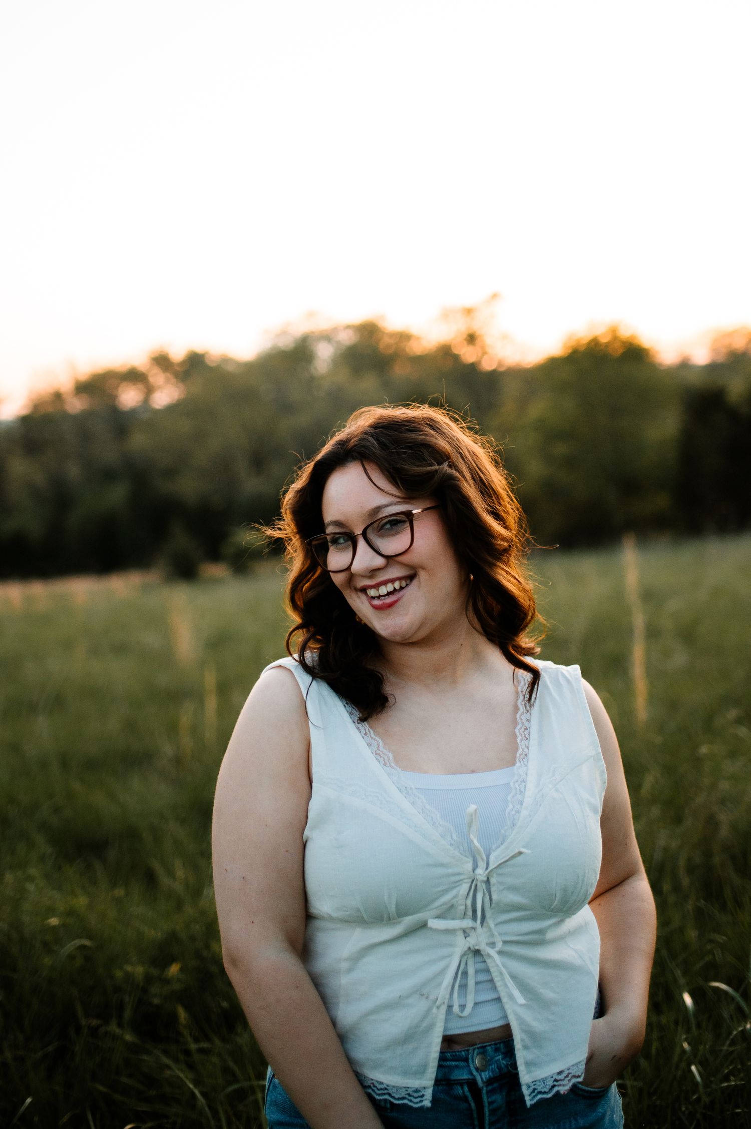 A person in glasses and white top smiles warmly during golden hour in a grassy field.