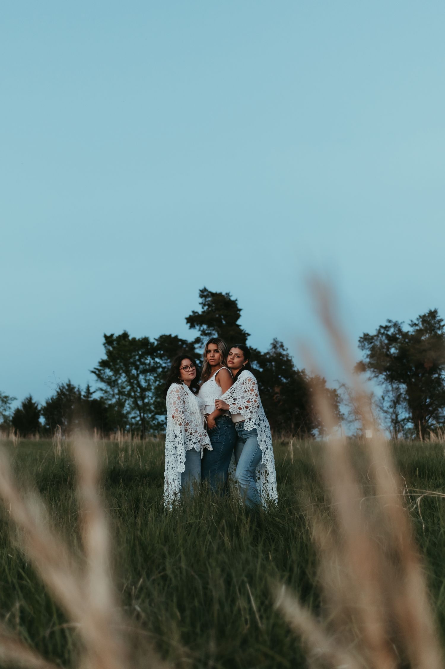 Figures in floral dresses stand together in a meadow during blue hour with trees in the background.