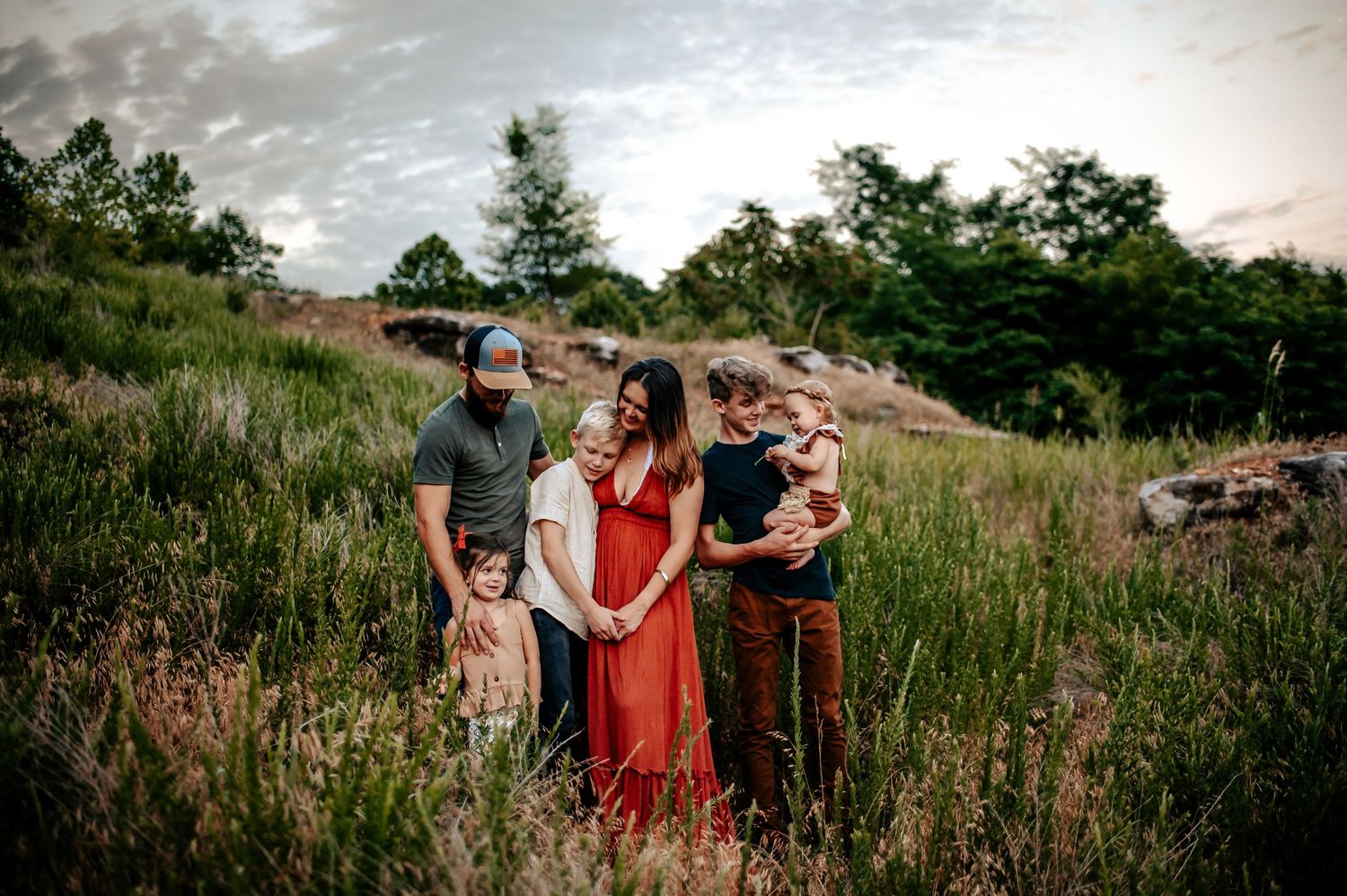 A family portrait in a meadow with tall grass and cloudy sky at sunset.