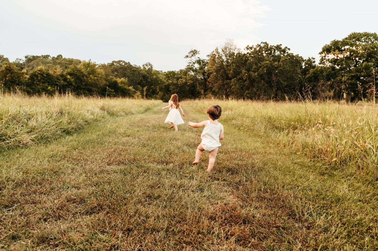 Two figures in white dresses running through a golden field at sunset with trees in the distance.