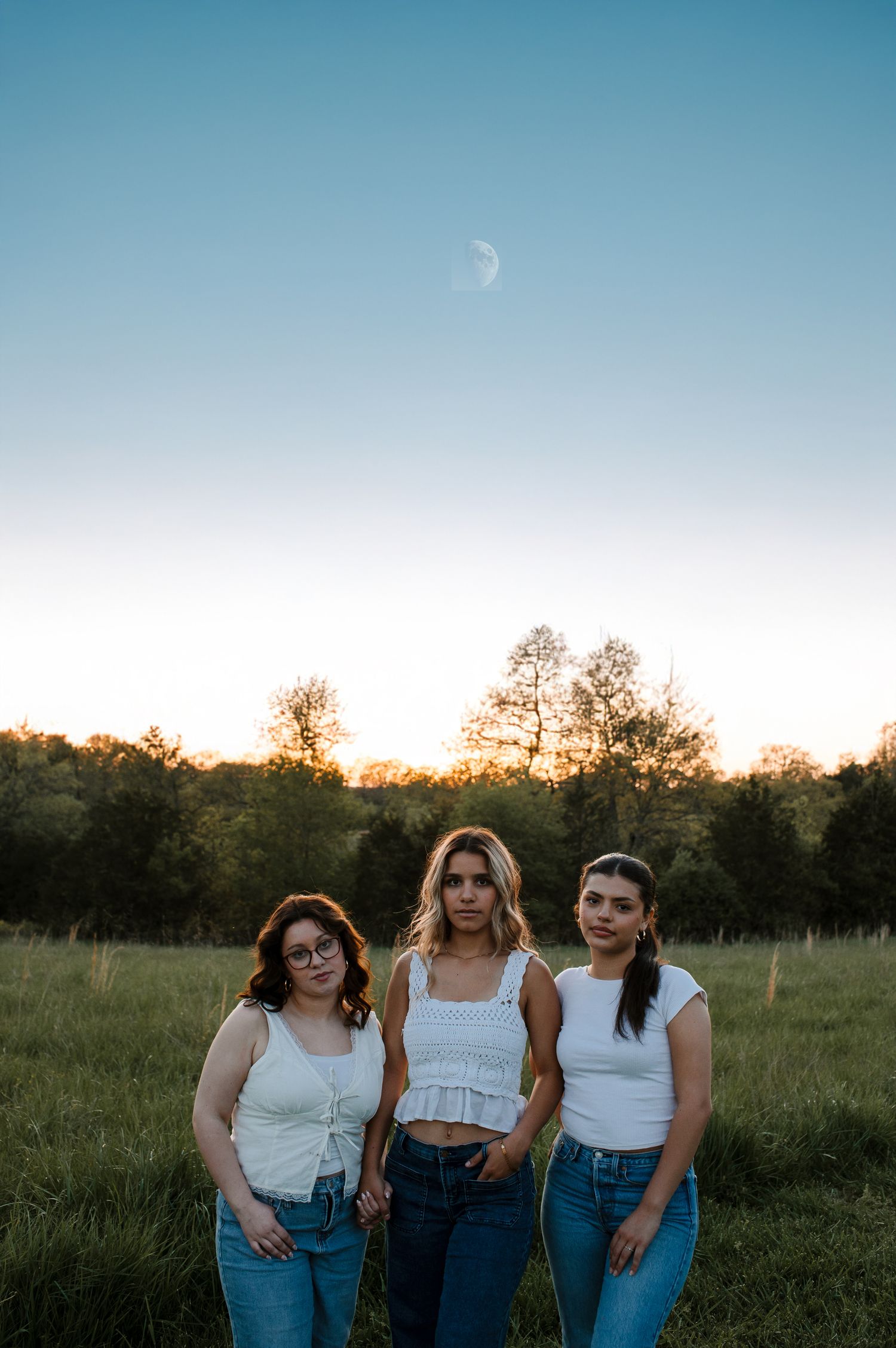 Three people in white tops and jeans stand together in a field at dusk with a crescent moon visible.