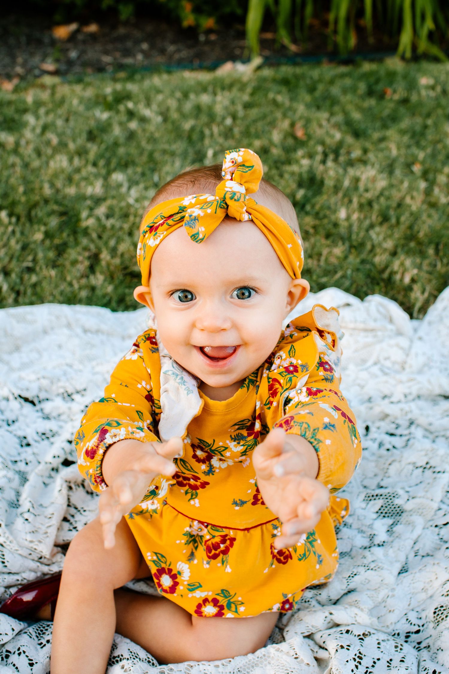 A smiling baby in a yellow floral dress and matching headband sitting on a white blanket.