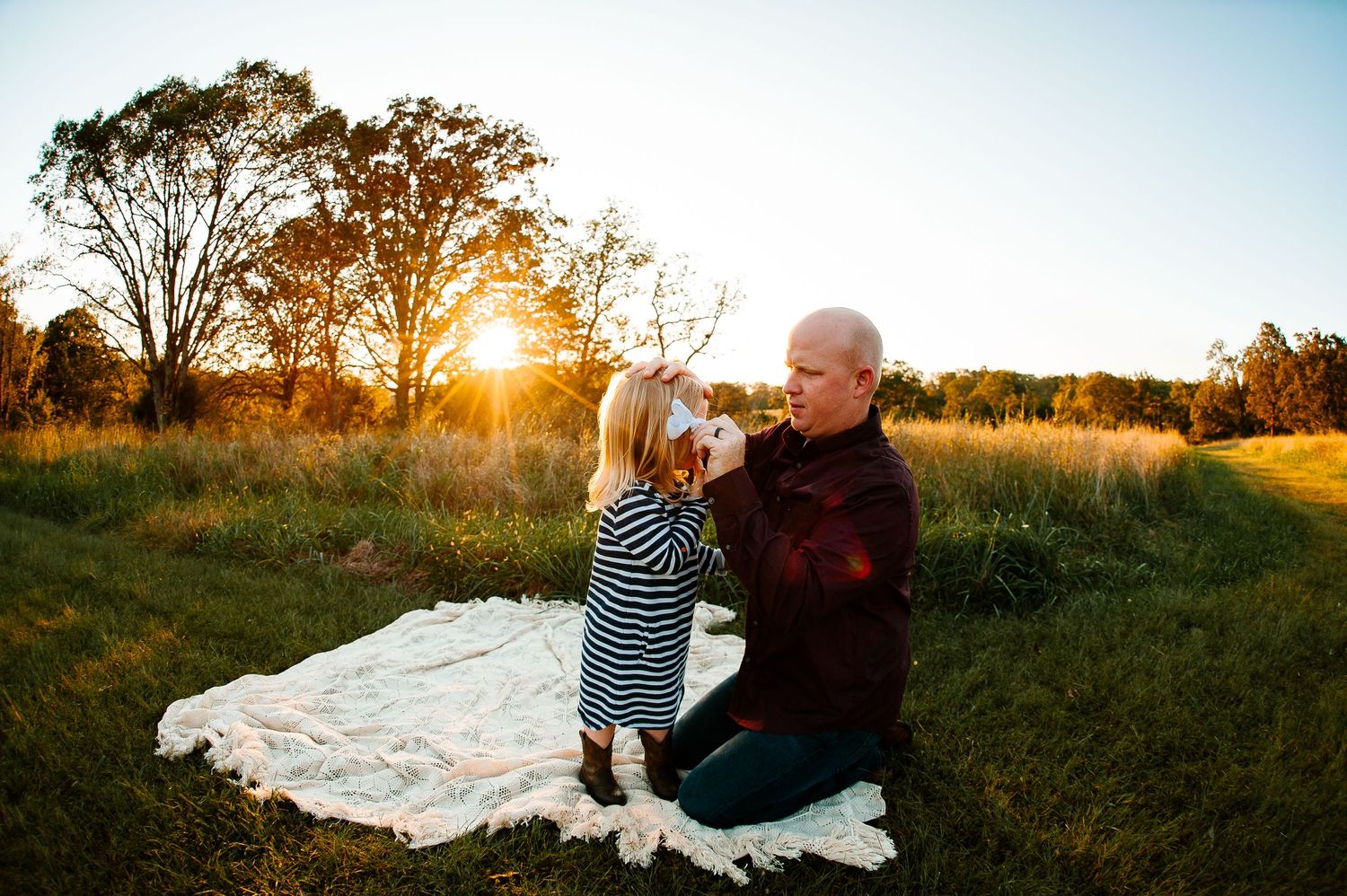 Sunset family moment on a white blanket in a grassy field with tall trees silhouetted in background.