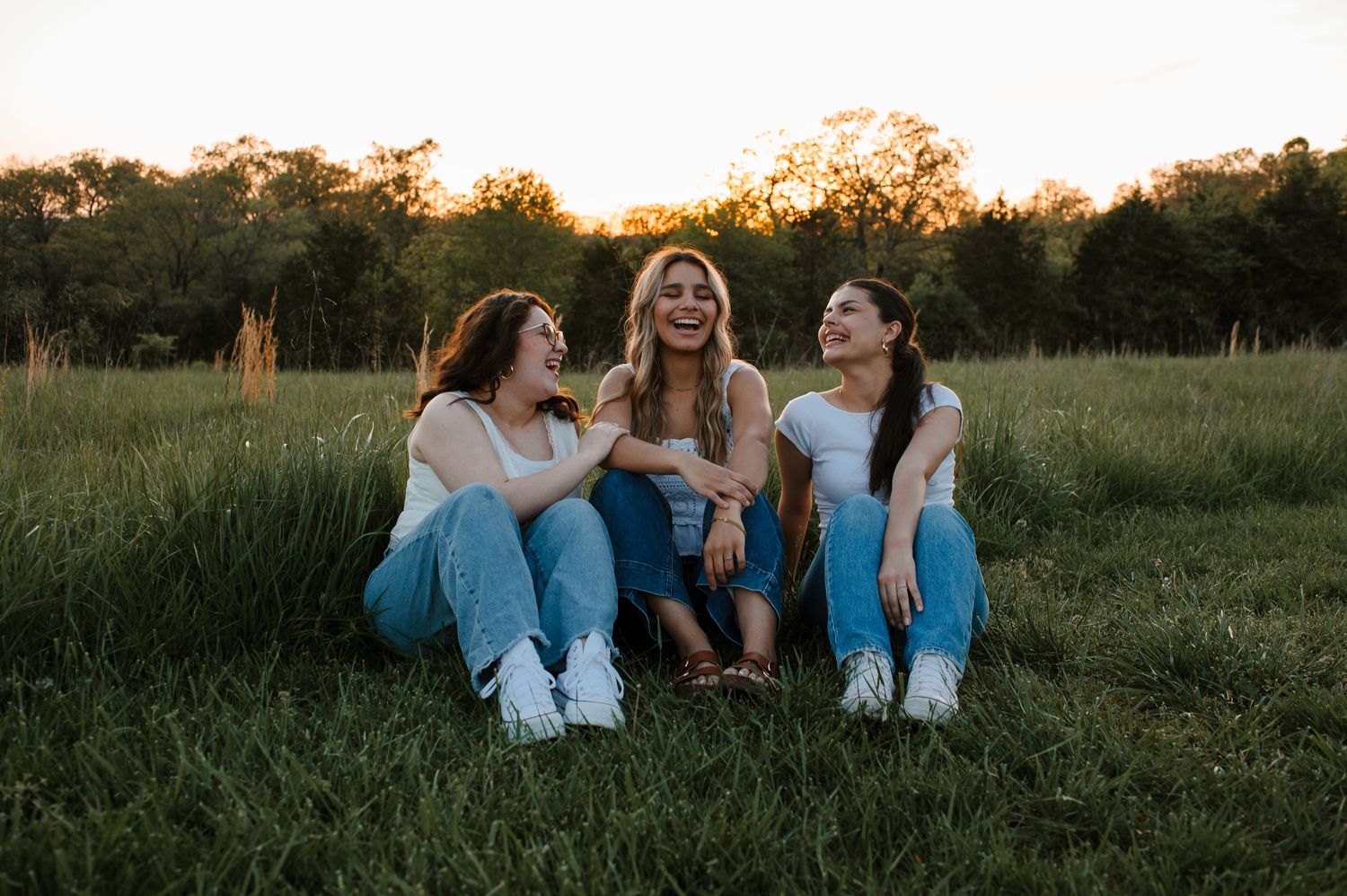 Three friends sitting together in a grassy field at sunset sharing laughs and conversation.