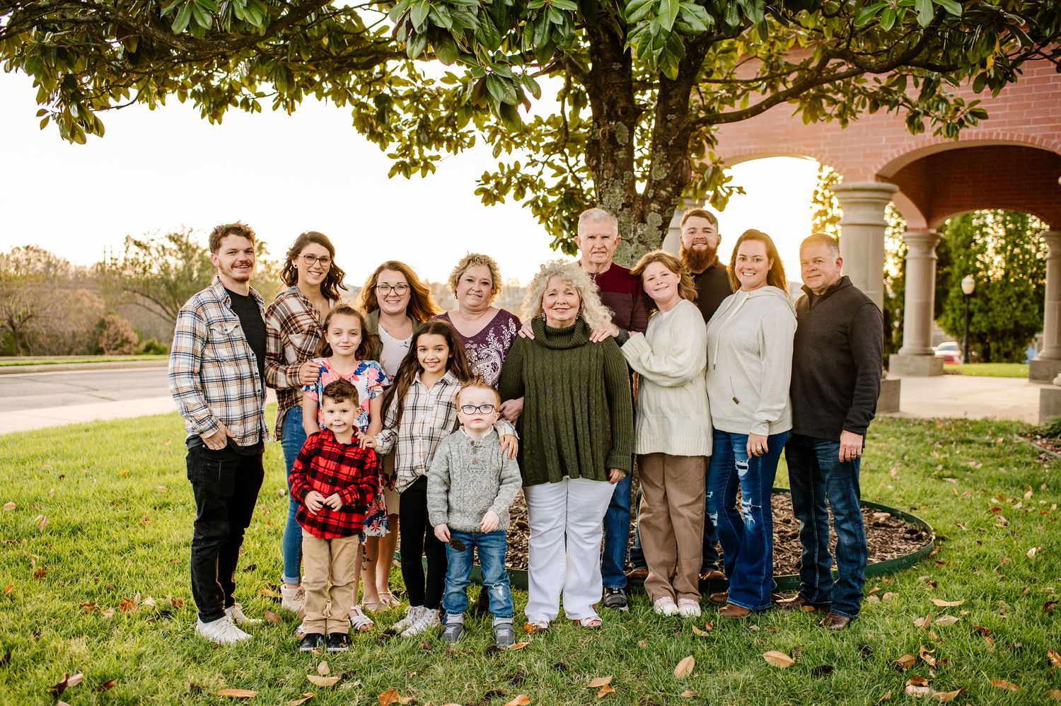 Large multigenerational family group portrait outdoors under a tree during autumn, smiling together on green grass.