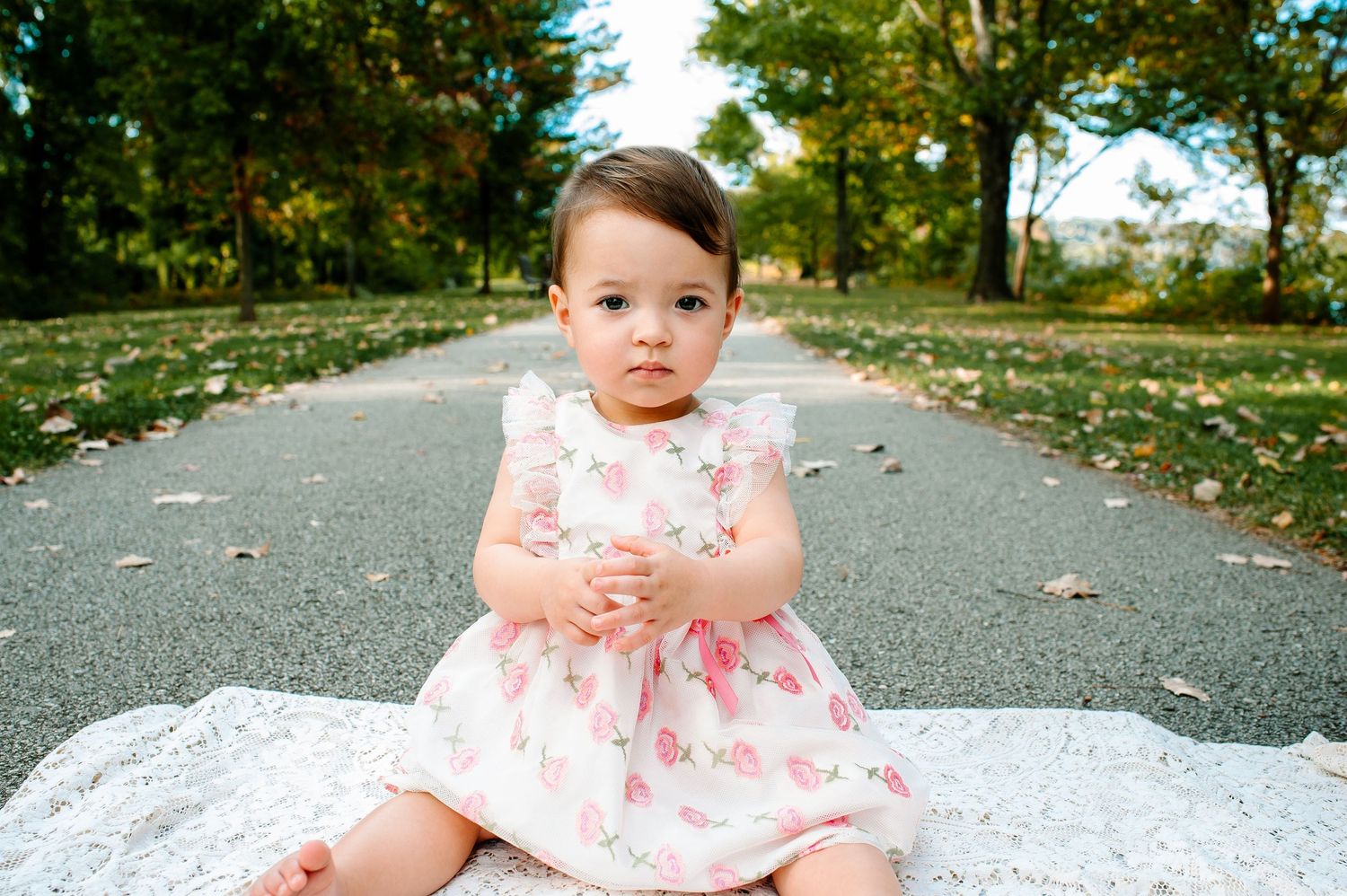 Baby in a floral dress sitting on a blanket on a leaf-covered path in autumn.