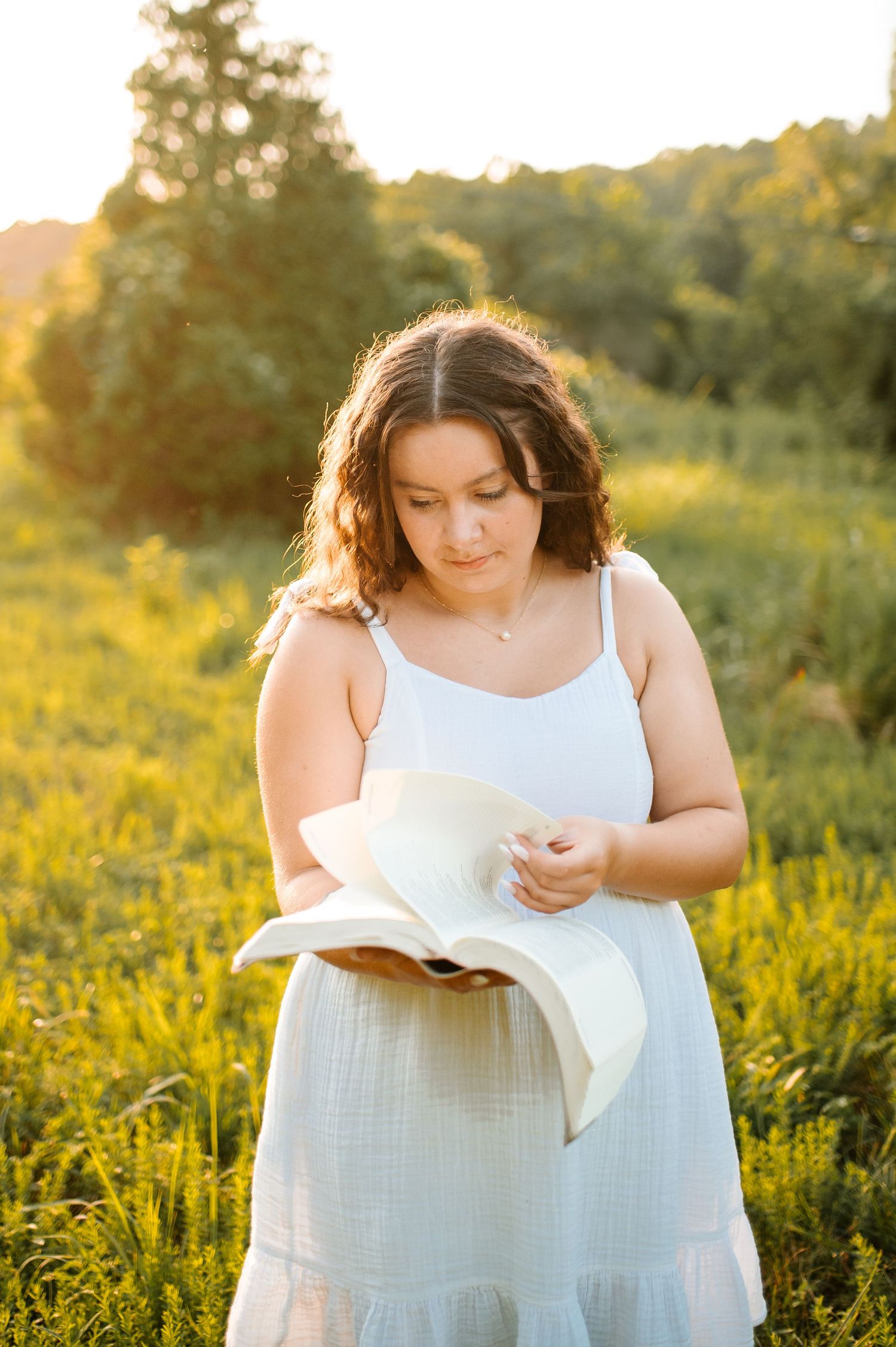 A person in a white dress reads a book while standing in a golden sunlit field of wildflowers at sunset.