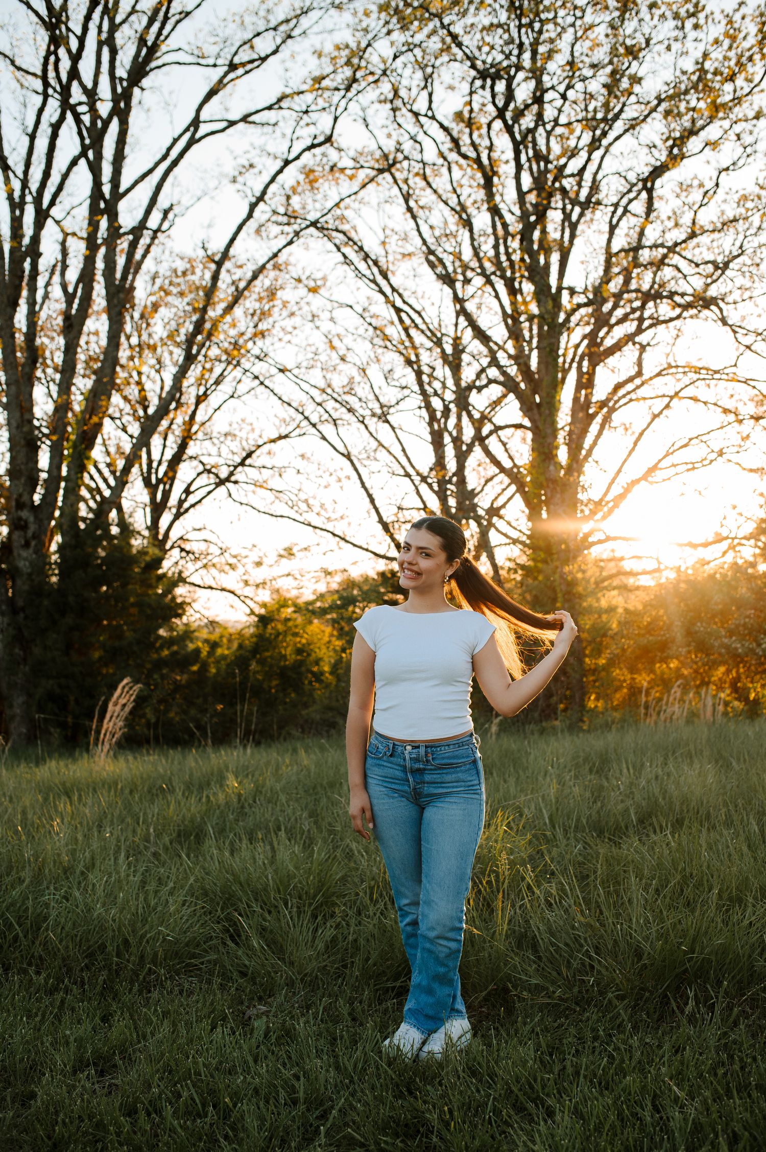 A person in a white top and jeans poses in a field at sunset with sunlight streaming through bare trees.