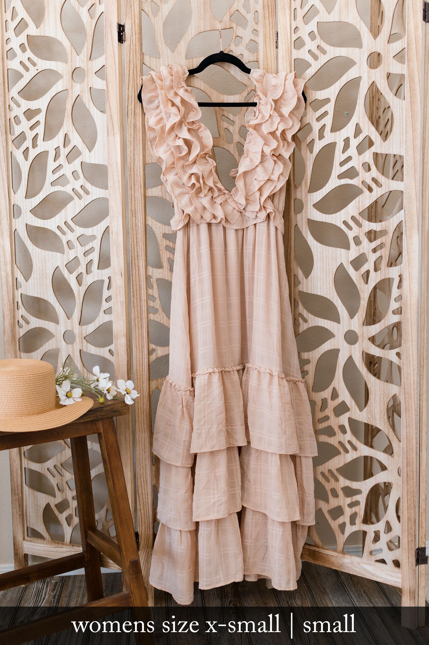 Feminine blush pink tiered maxi dress with ruffled neckline hanging on display against decorative white floral wall panel.