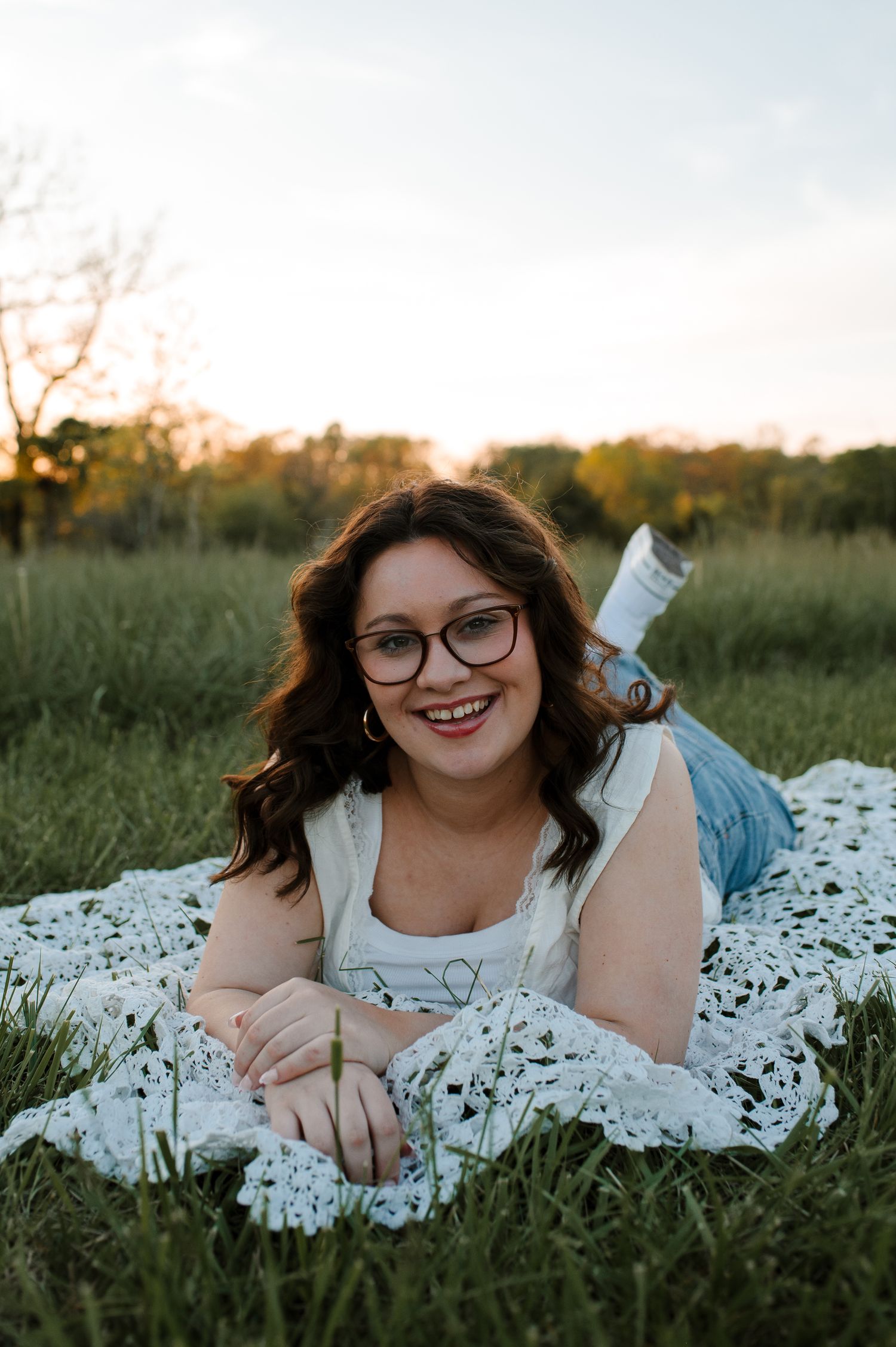 A person in glasses lies on a white blanket in a grassy field during golden hour.