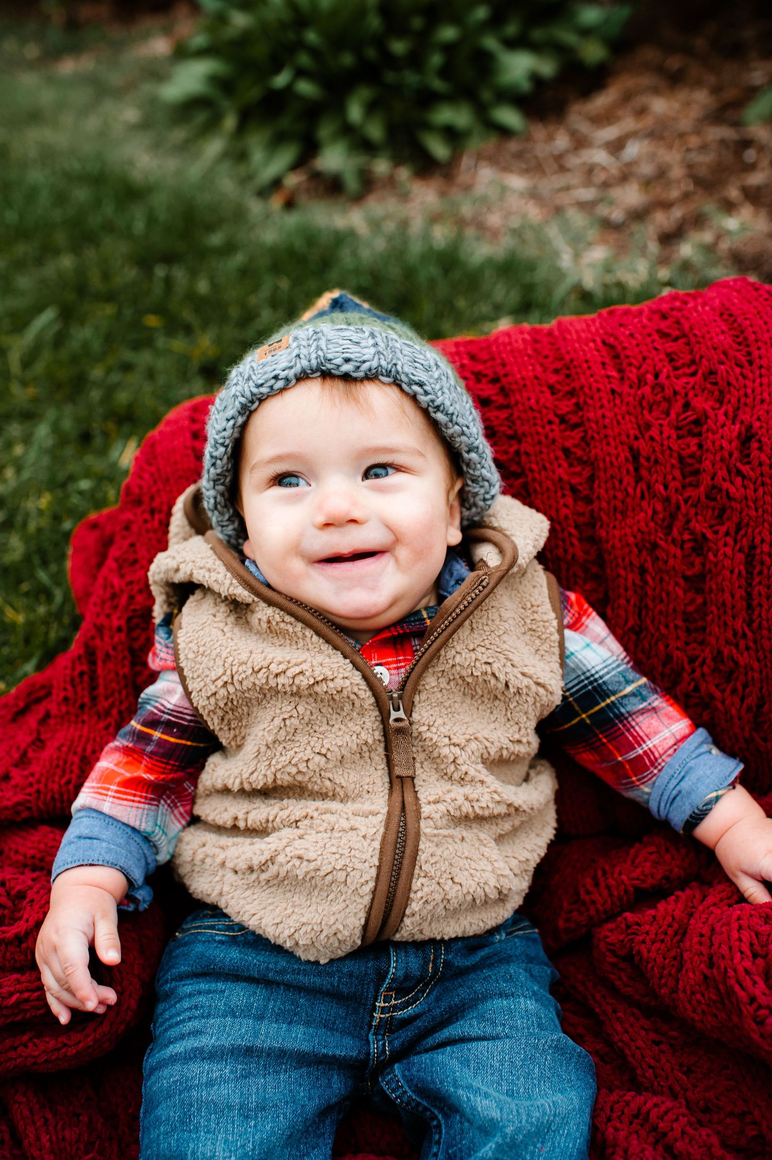 A smiling baby wearing a gray knit hat and fuzzy vest sits on a red blanket outdoors.