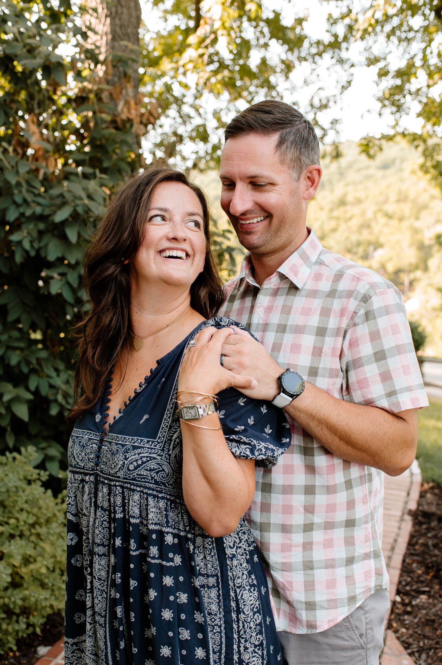 A couple shares a joyful moment together outdoors, laughing in warm sunlight with trees and foliage in the background.