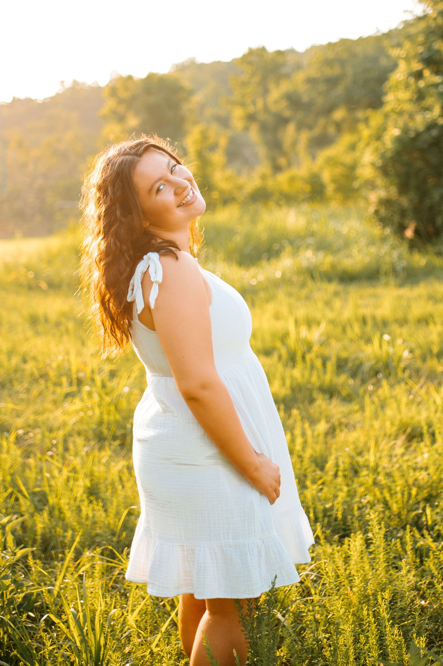 A glowing sunset illuminates a grassy meadow as someone in a white summer dress poses during golden hour.