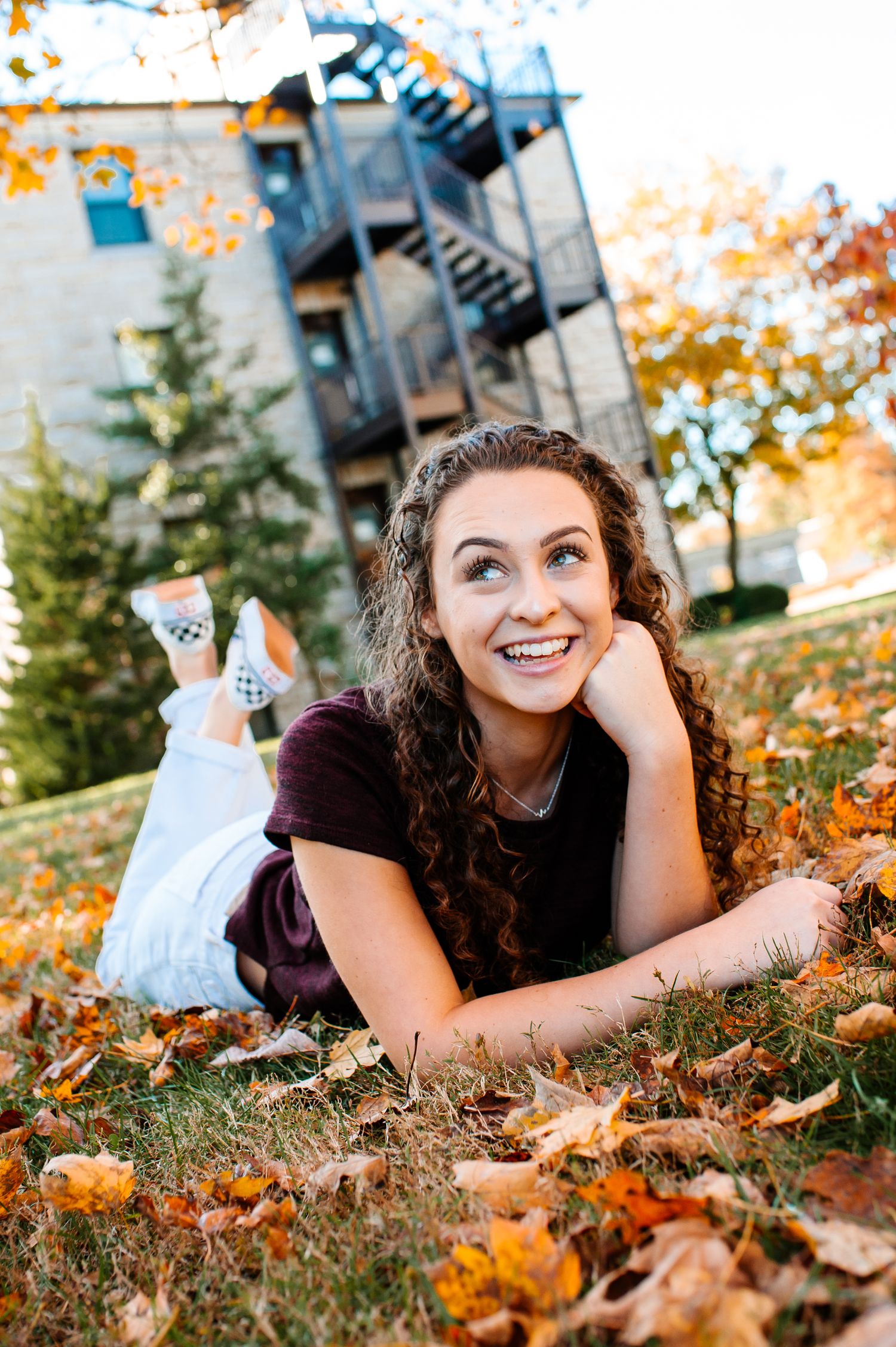 Someone with curly hair lies among fall leaves wearing a black shirt and patterned pants.