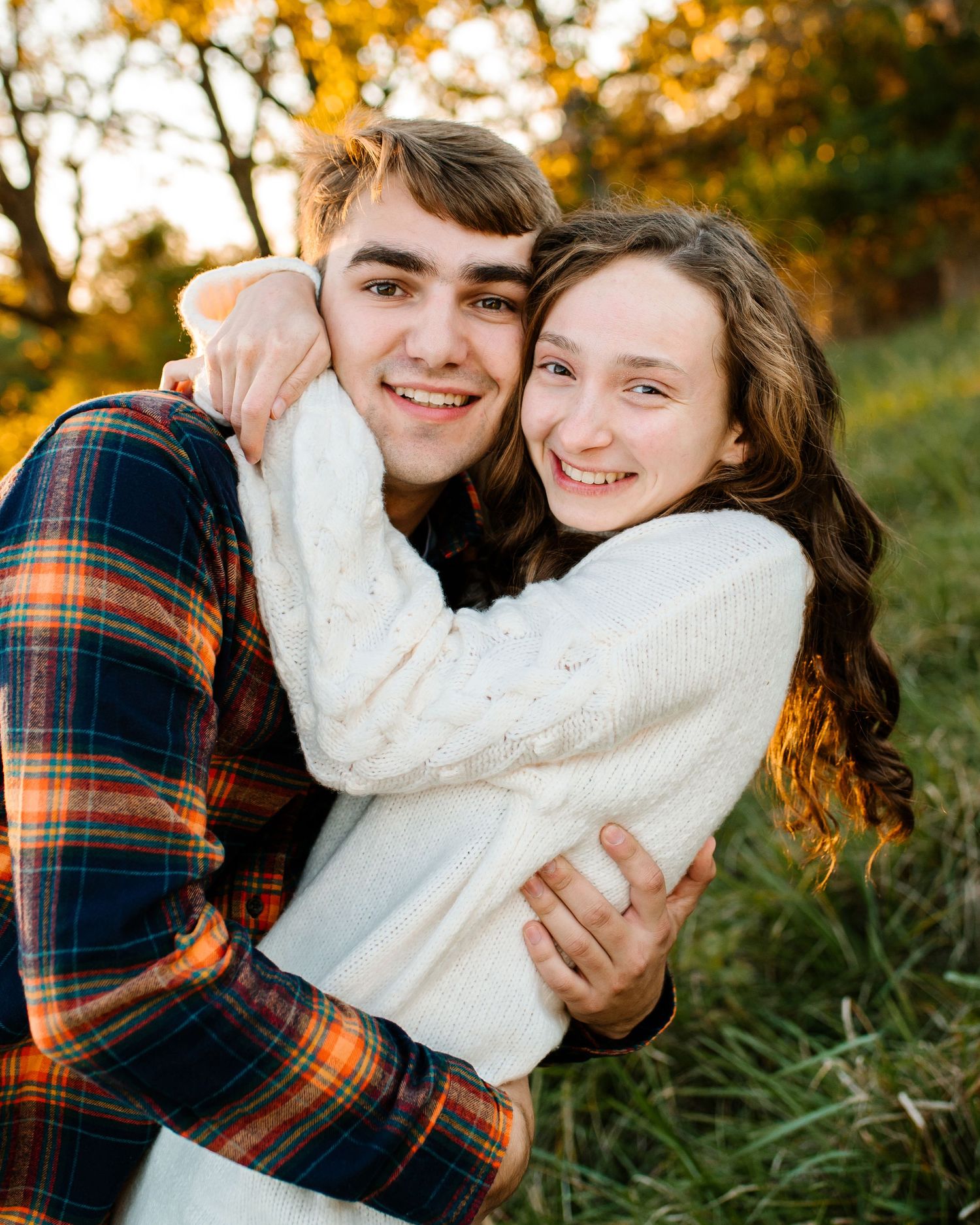 A couple in casual fall attire embraces and smiles during golden hour in a grassy field.