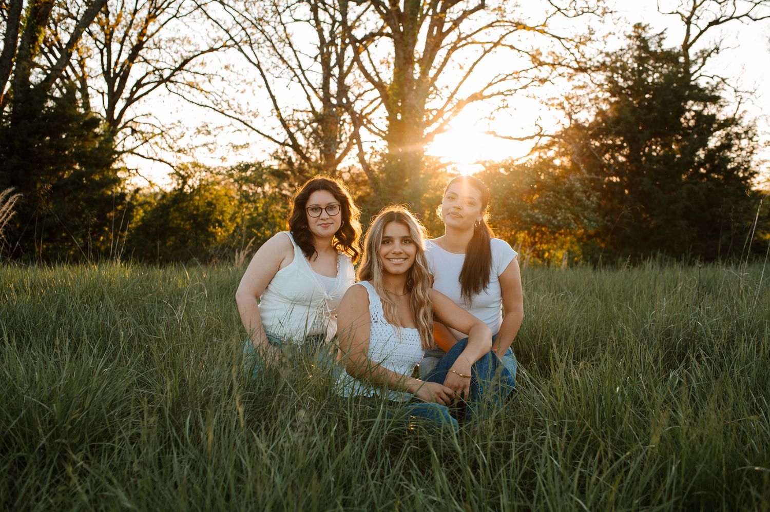 Three people in white tops sit together in a grassy field at sunset with golden light filtering through trees.