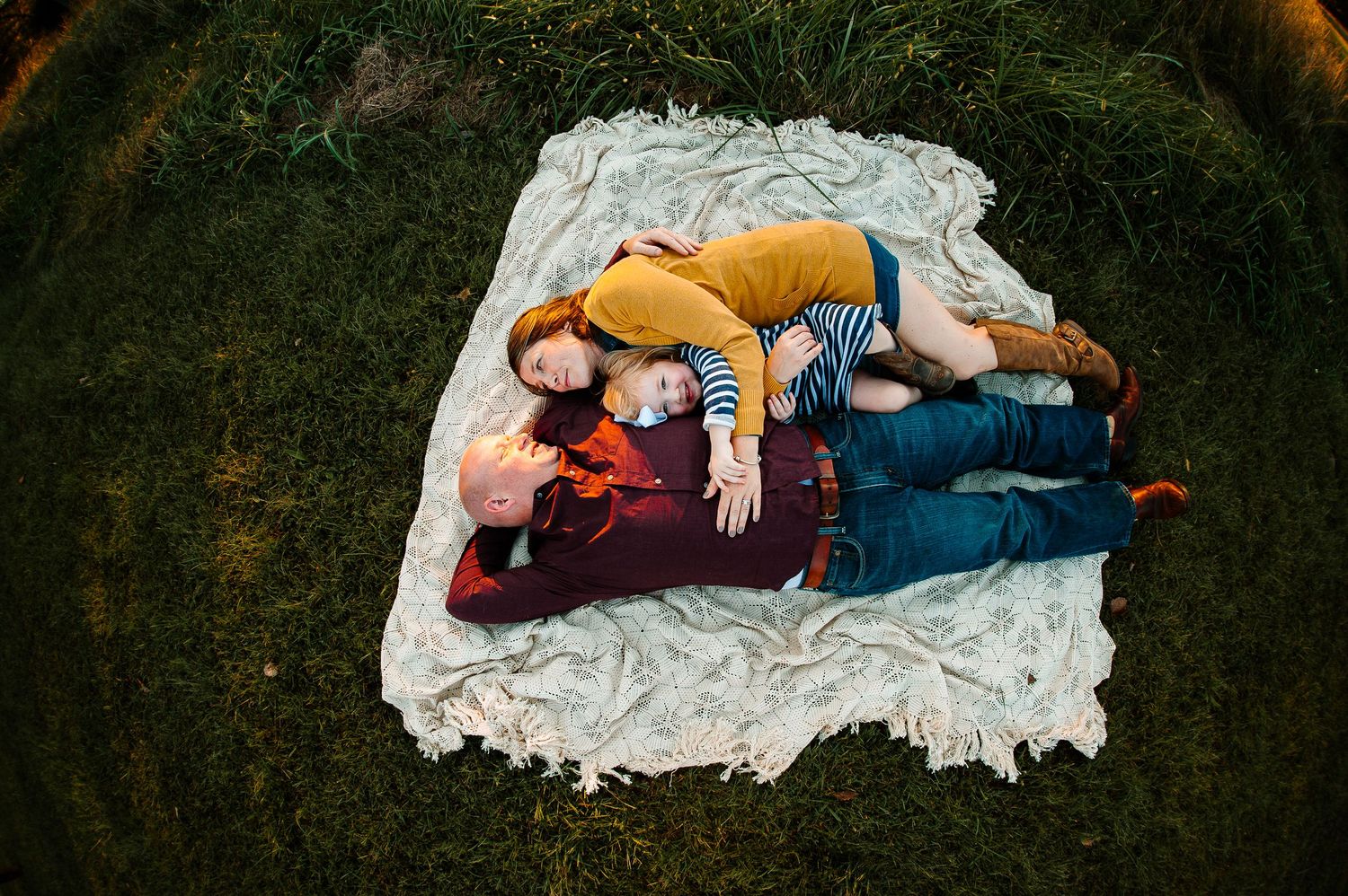 Overhead view of a family lying together on a white blanket in autumn grass during golden hour.