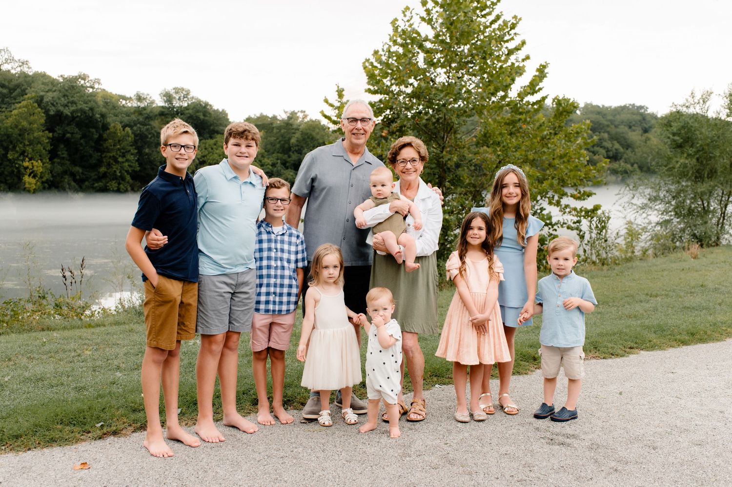 Multi-generational family portrait taken outdoors by a lake with trees in the background.