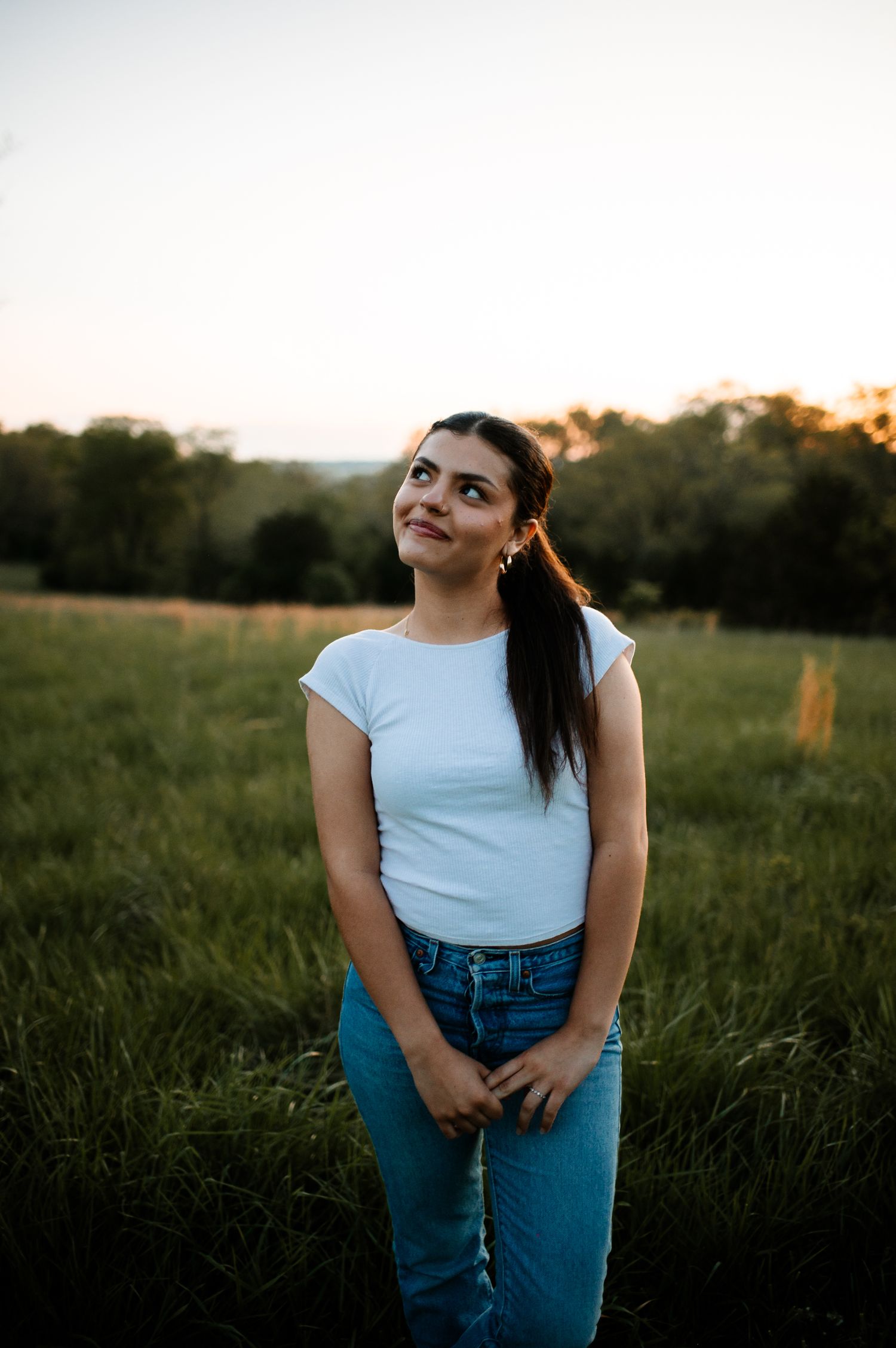 Person in white t-shirt and jeans poses playfully in a sunlit meadow at dusk.