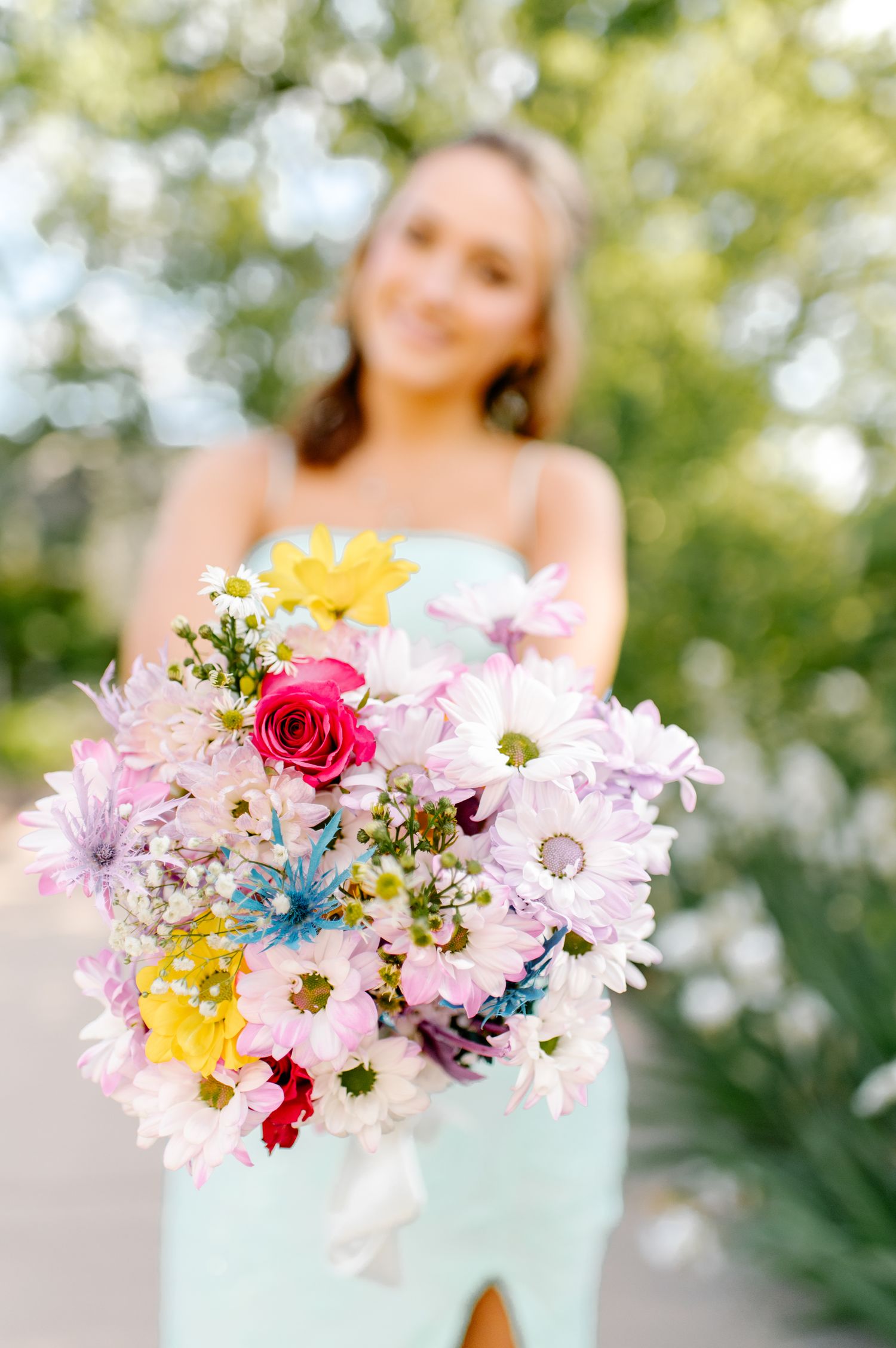 Colorful spring bouquet with pink daisies, roses and blue accents in a mint green vase against a soft blurred background.