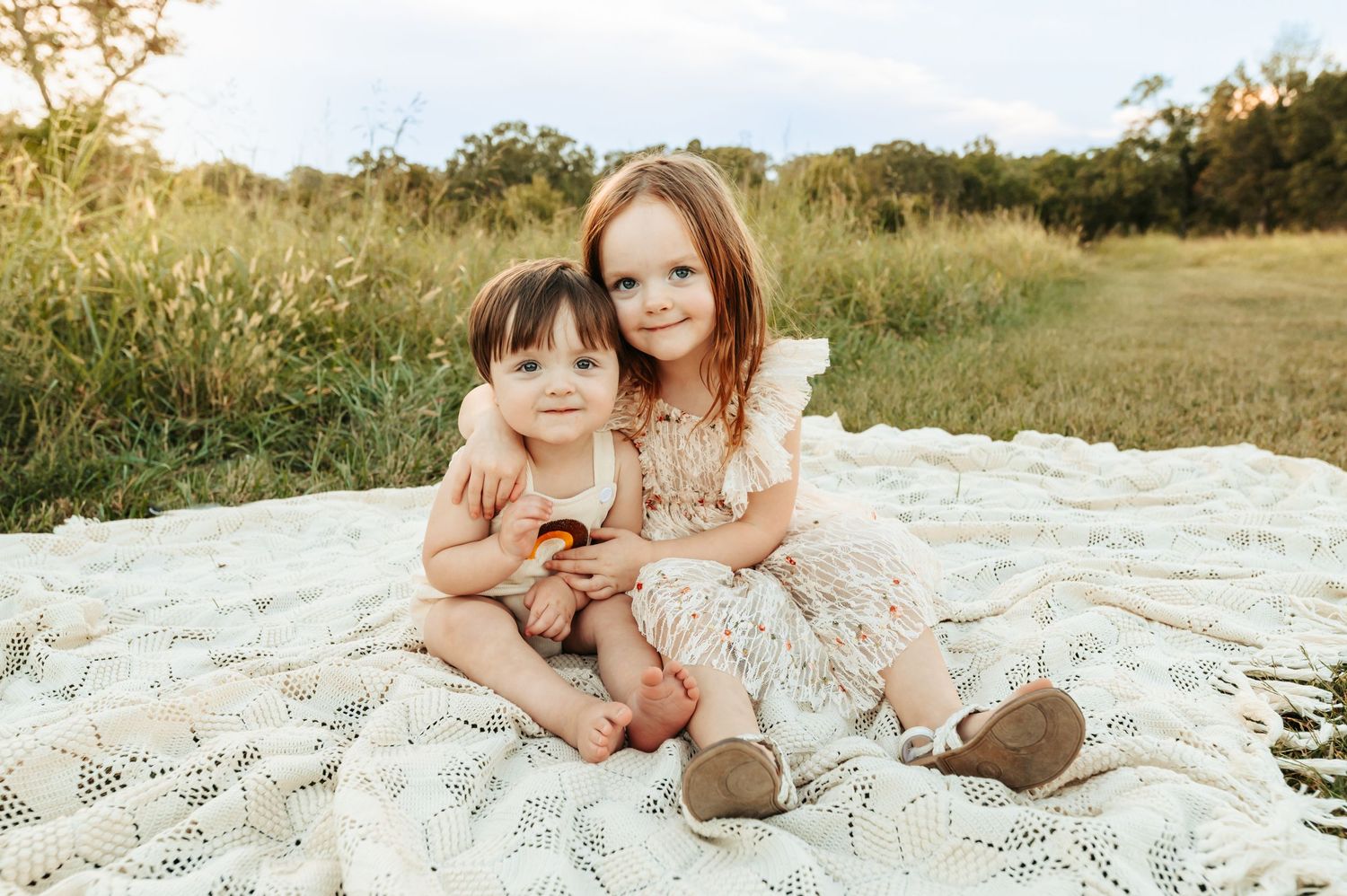 Two young children sitting on a white blanket in a grassy field during sunset.