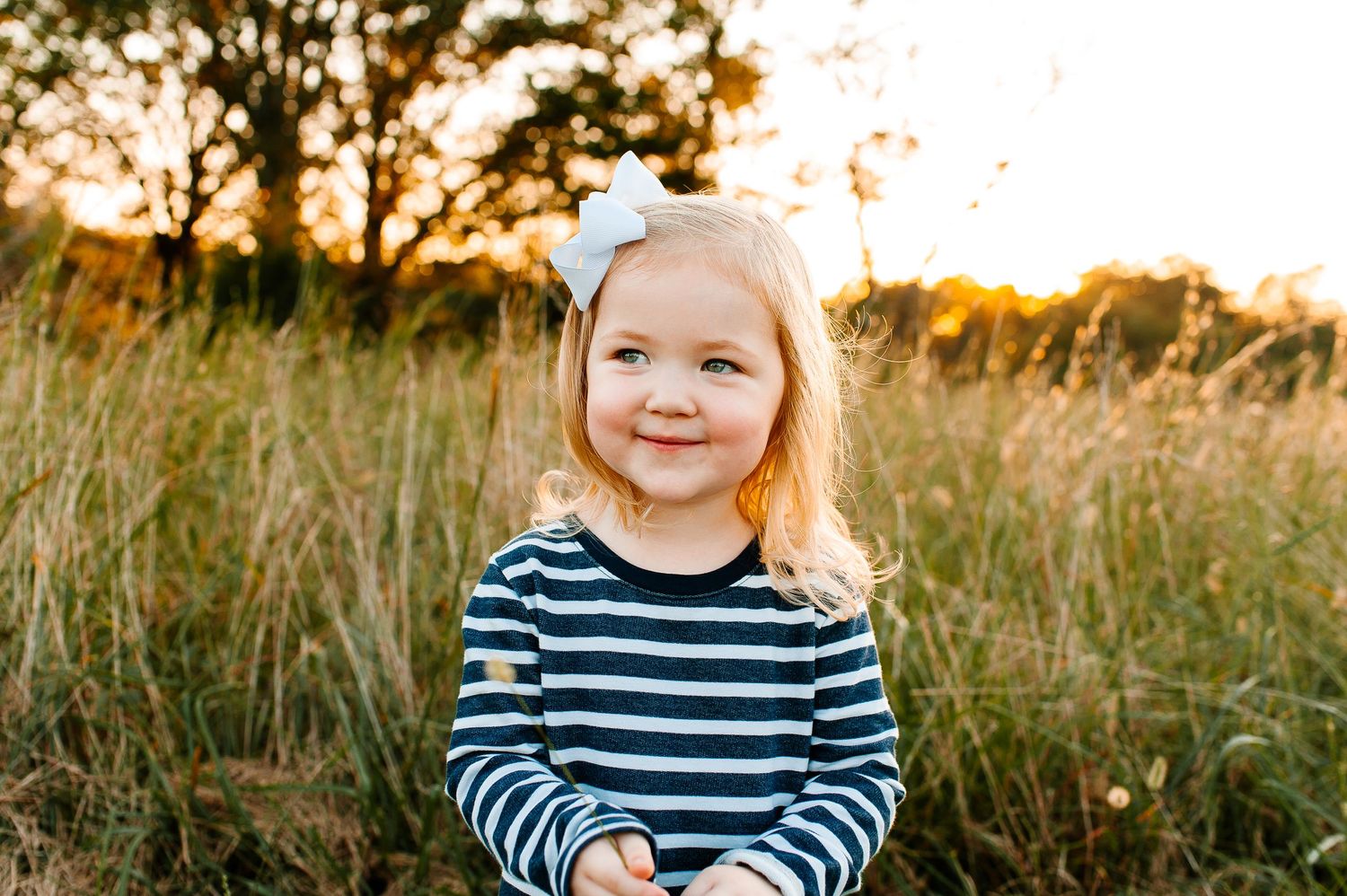 Child in navy striped shirt smiling outdoors at golden hour with tall grass in background.