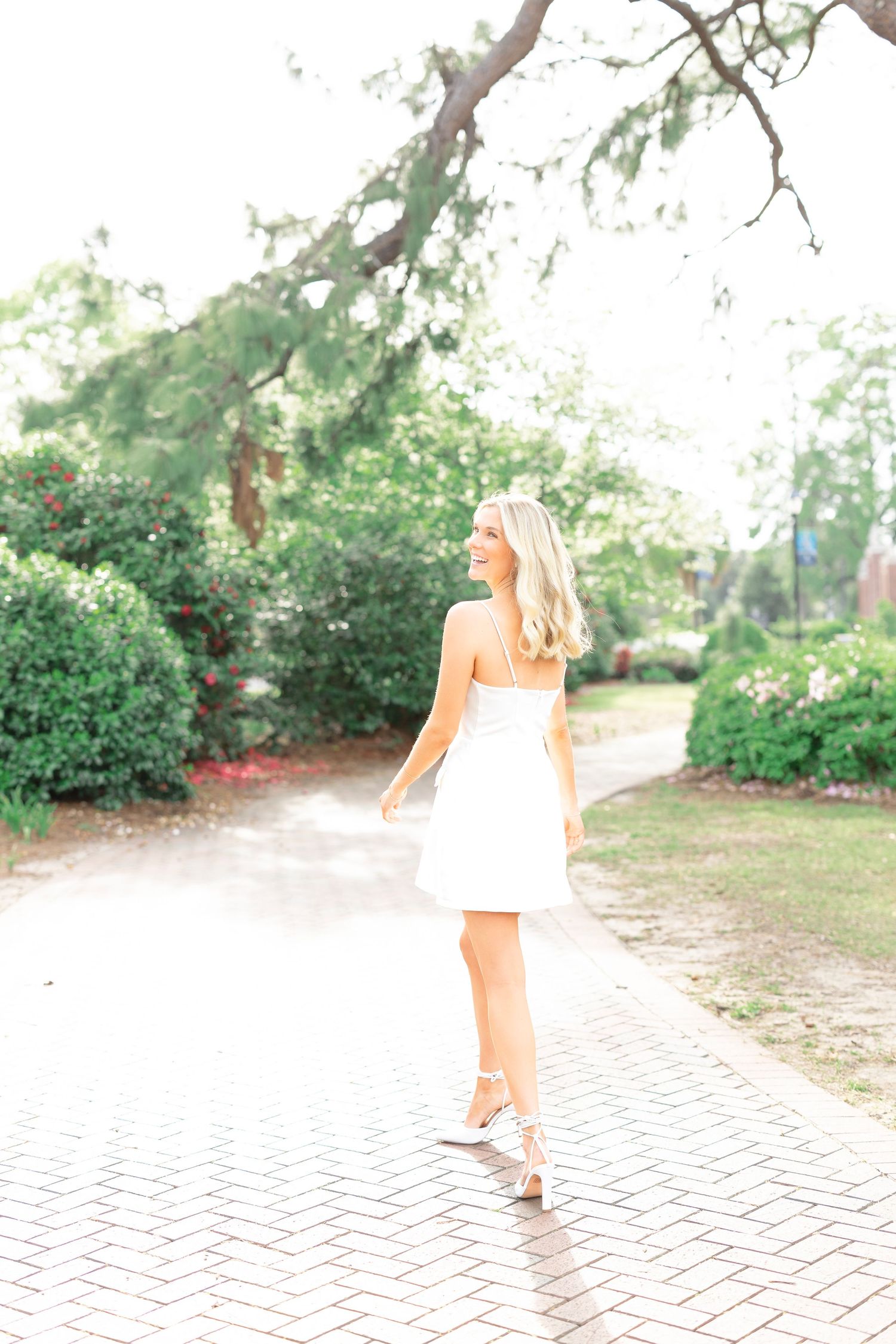 A woman in a white dress walks along a brick pathway in a garden setting.