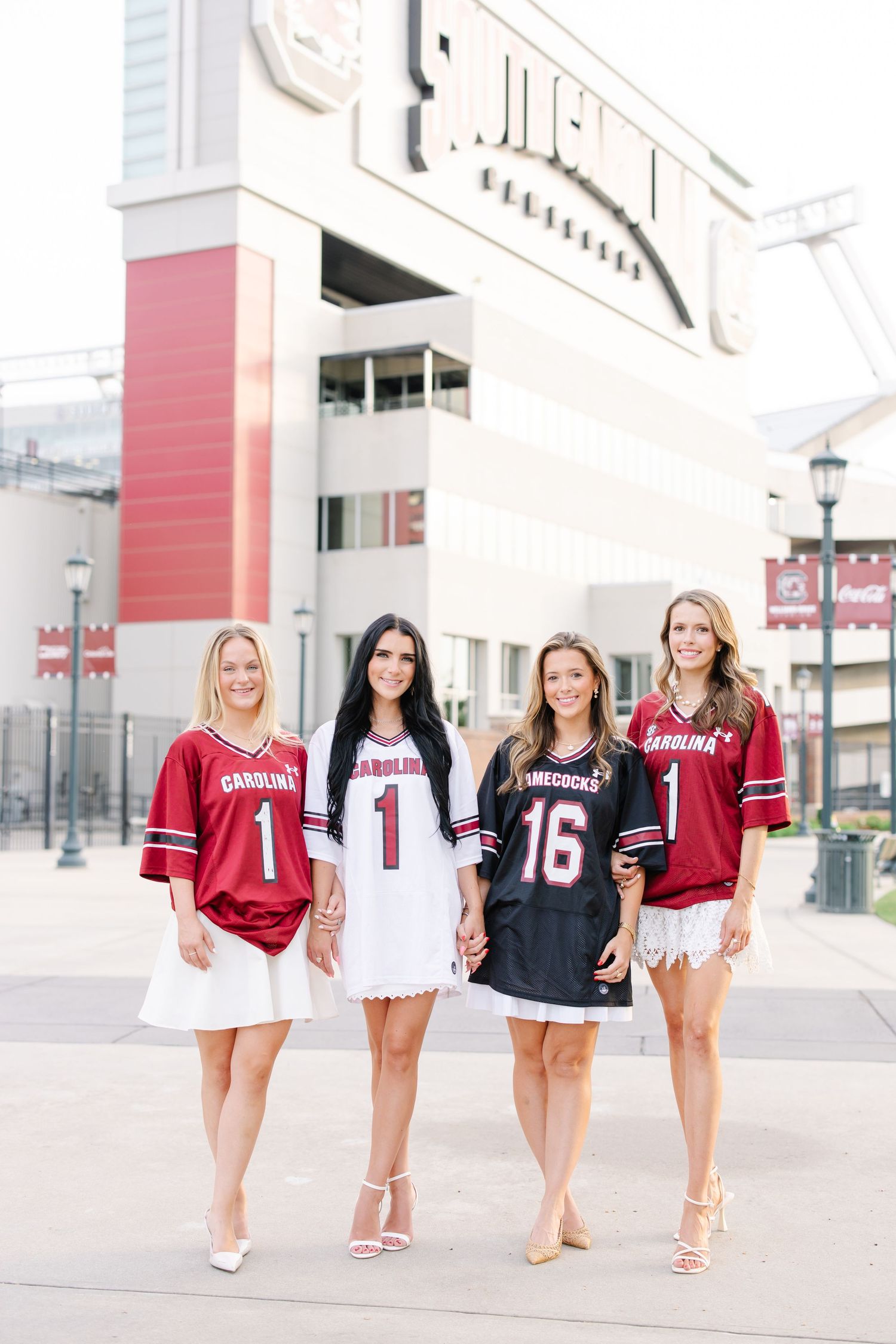 Four women wearing sports jerseys pose together on a walkway outside a stadium.